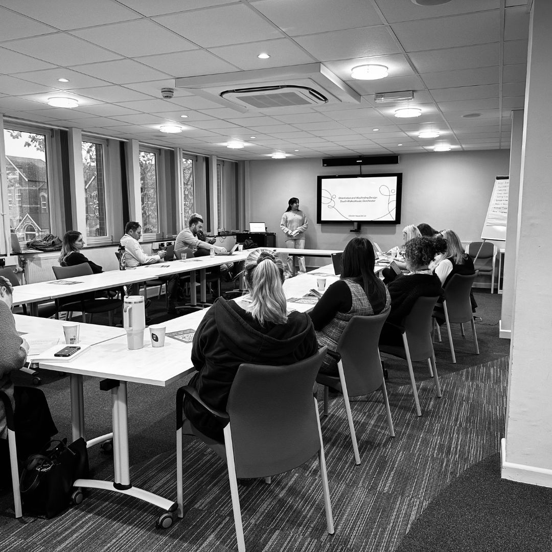 A person giving a pitch presentation to a group of people sat around a quadrangle of tables.