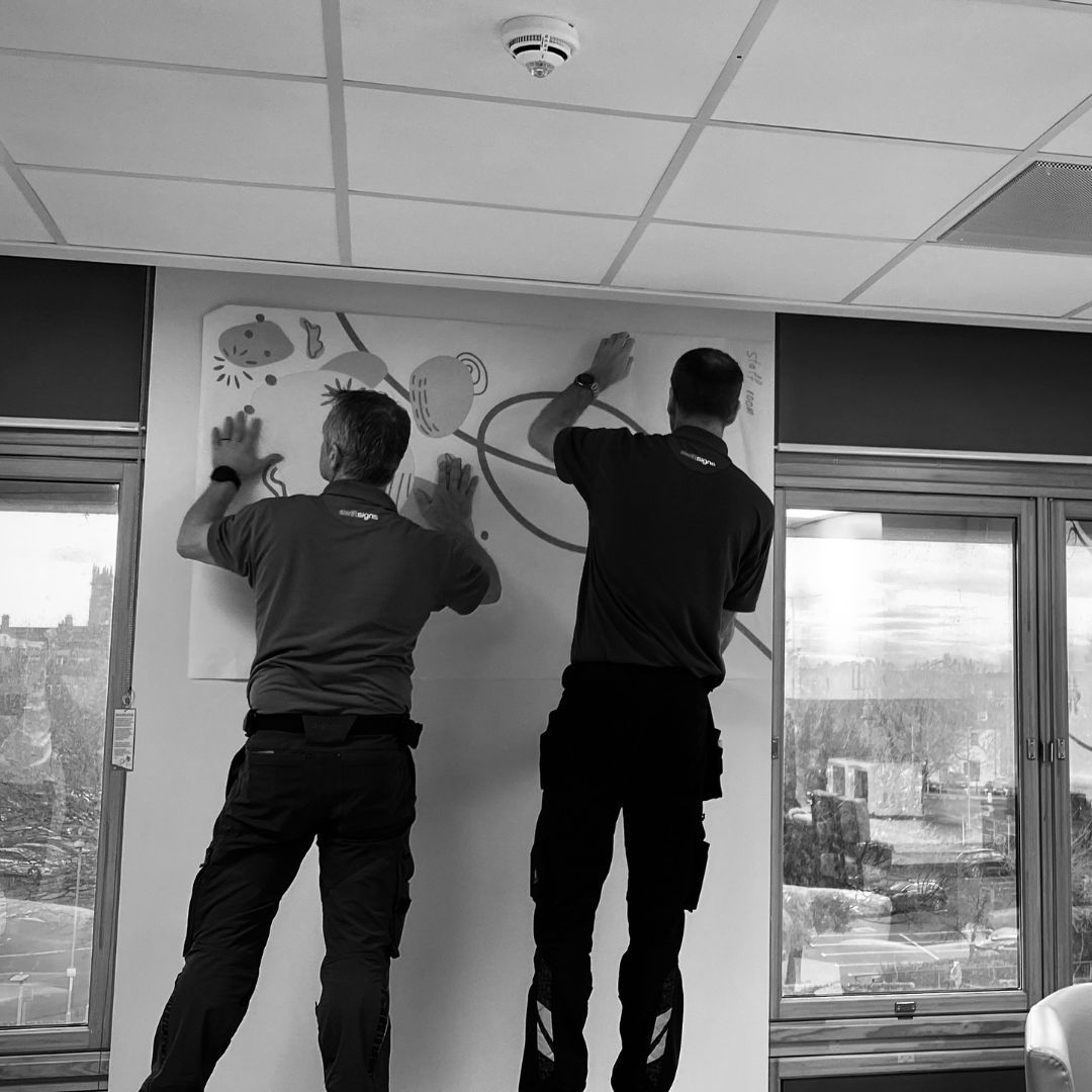 Two men viewed from behind putting up a piece of artwork with an abstract design on a wall.