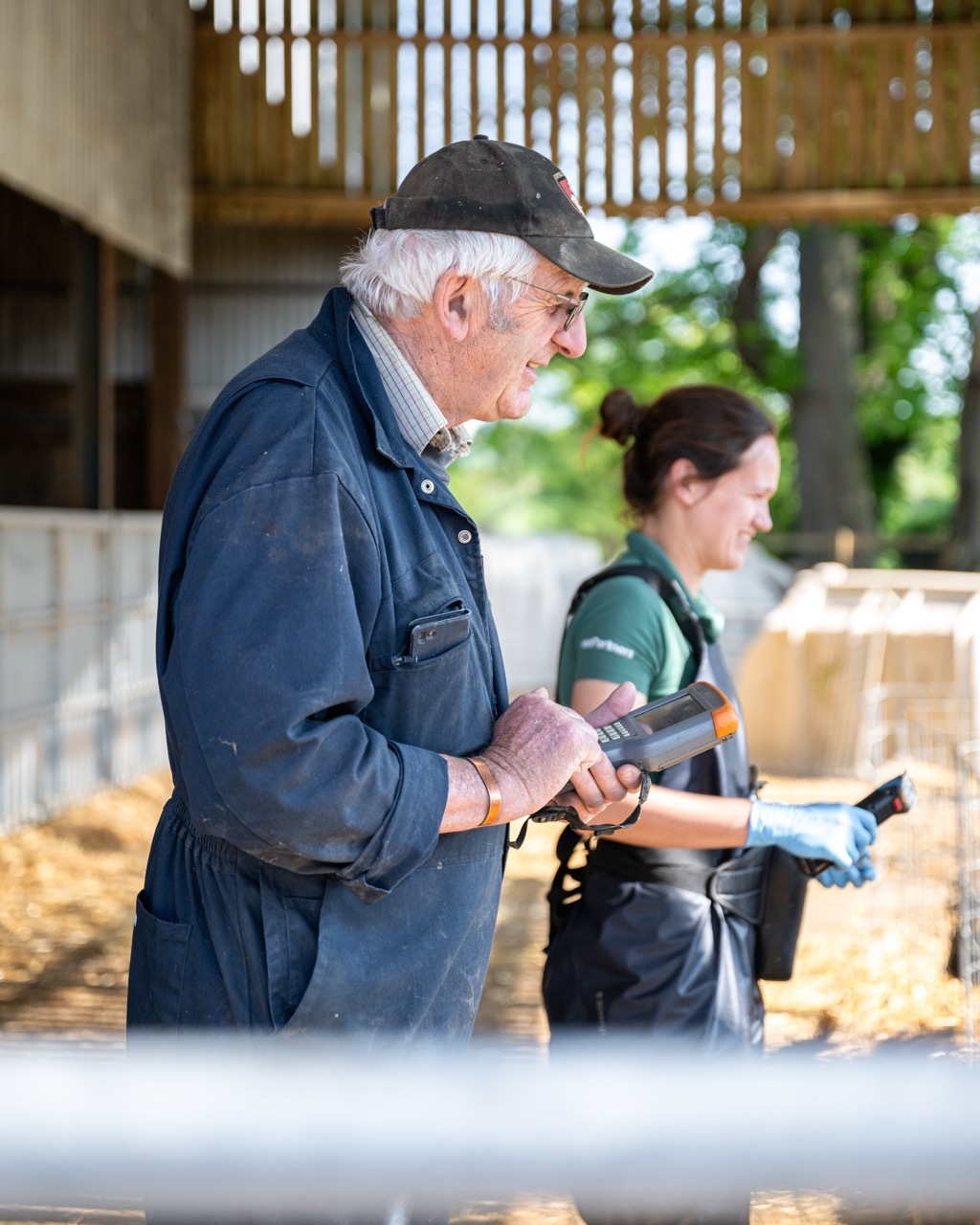 Elderly man in dark cap and jacket using device, woman in green top working behind him in outdoor workshop setting.