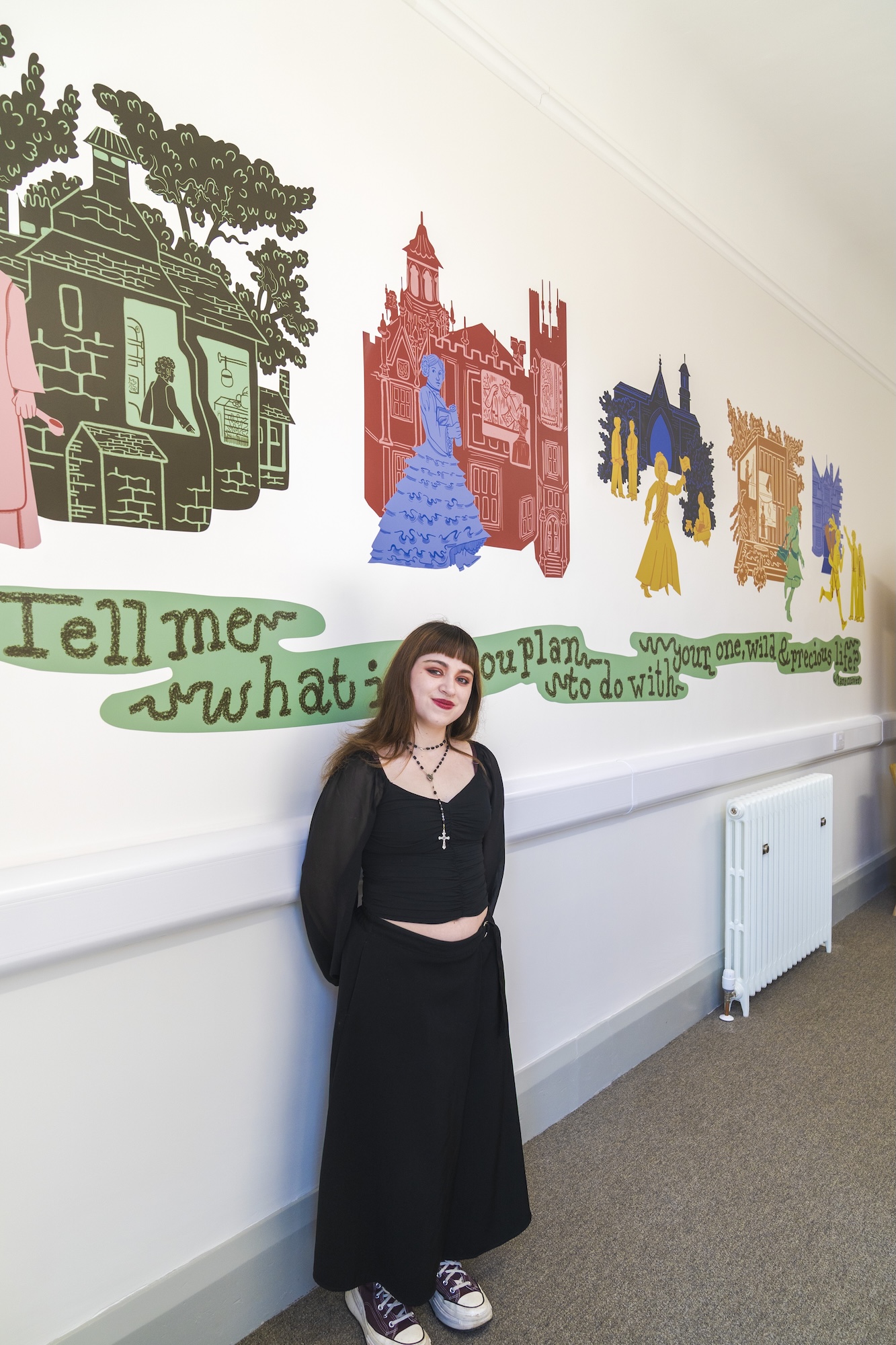 BA (Hons) Illustration student R Sansores posing for a photo in front of a series of colourful vinyl illustrations of houses on the wall behind them.
