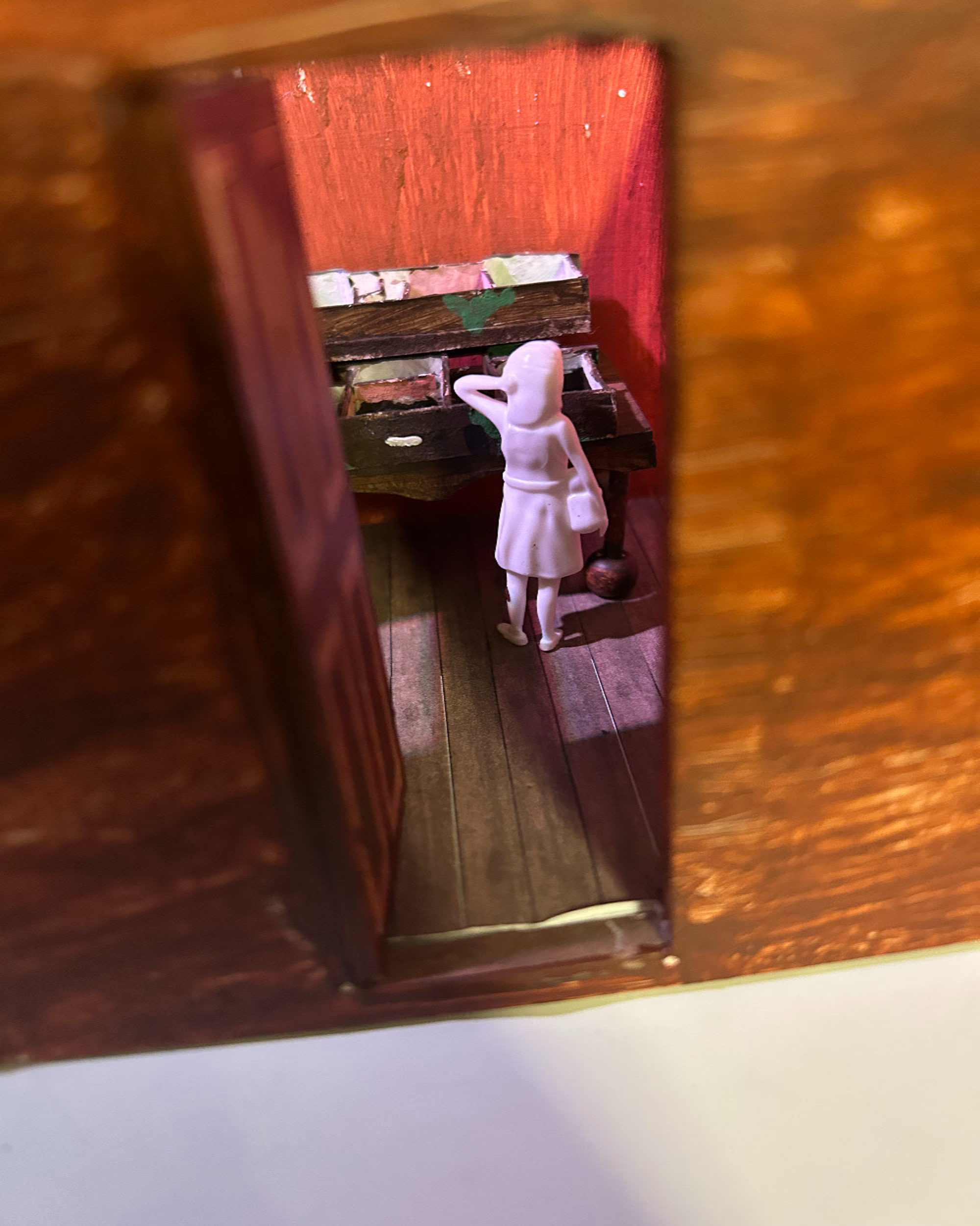 White figure seated at piano inside miniature red-walled dollhouse room, viewed through doorway opening on wooden surface.