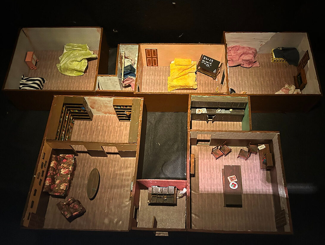 Overhead view of dollhouse with multiple rooms containing miniature furniture, coloured objects, and striped wallpaper in cardboard compartments.