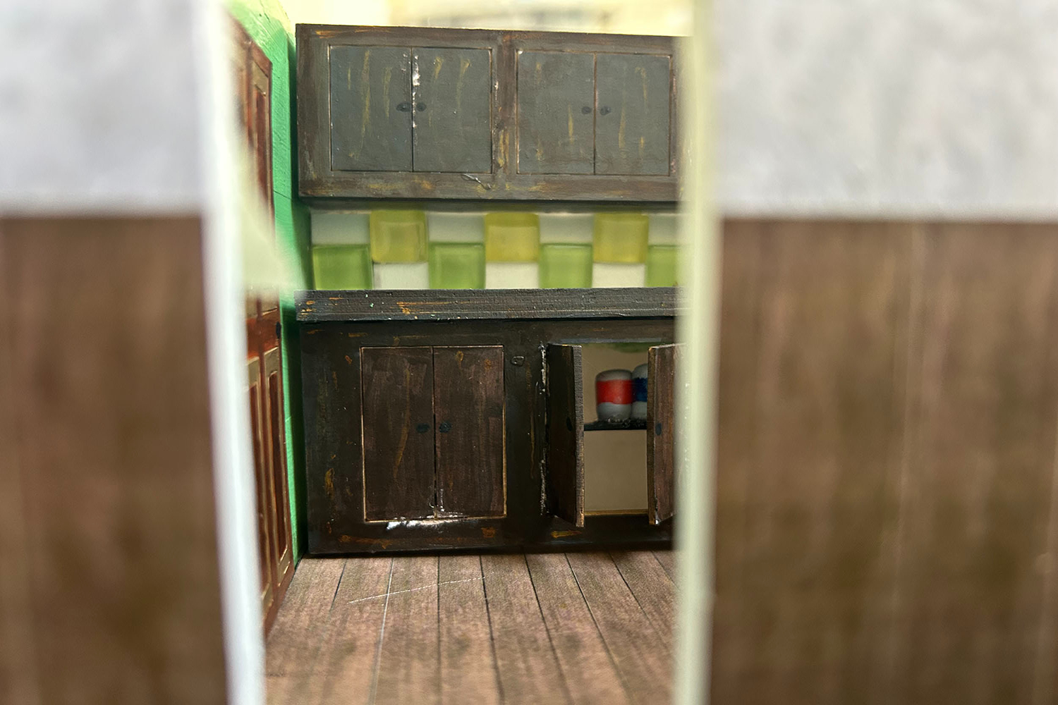Miniature kitchen with dark wooden cabinets, yellow-green backsplash tiles, and wooden flooring viewed through white doorframe.