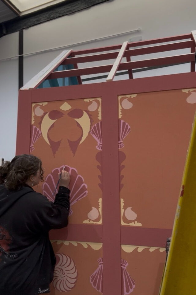 Person painting burgundy wall mural with decorative shell and fan motifs in pink, cream, and gold colours beneath white staircase railing.