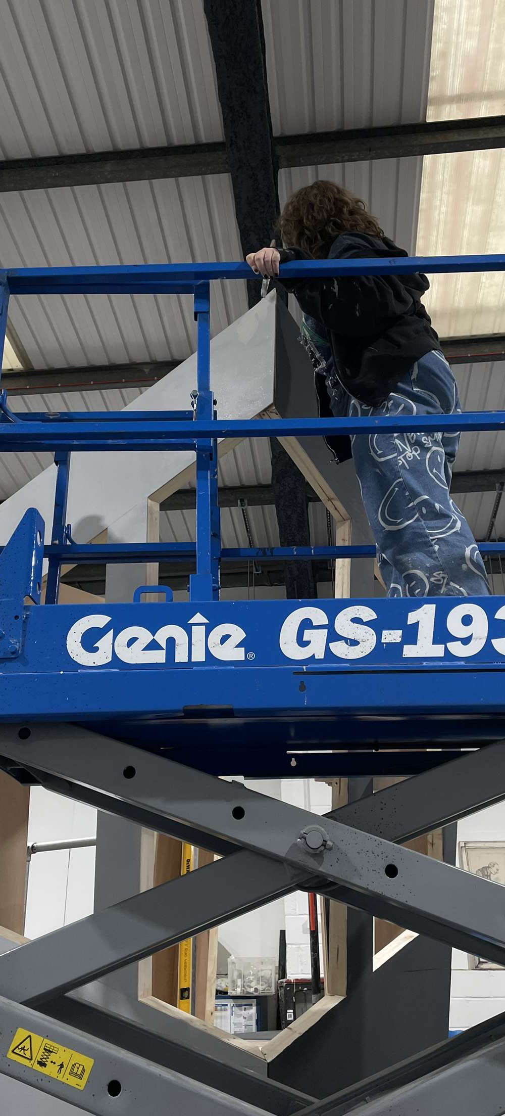 Person on blue Genie GS-19 scissor lift working inside warehouse with corrugated metal ceiling and steel framework.