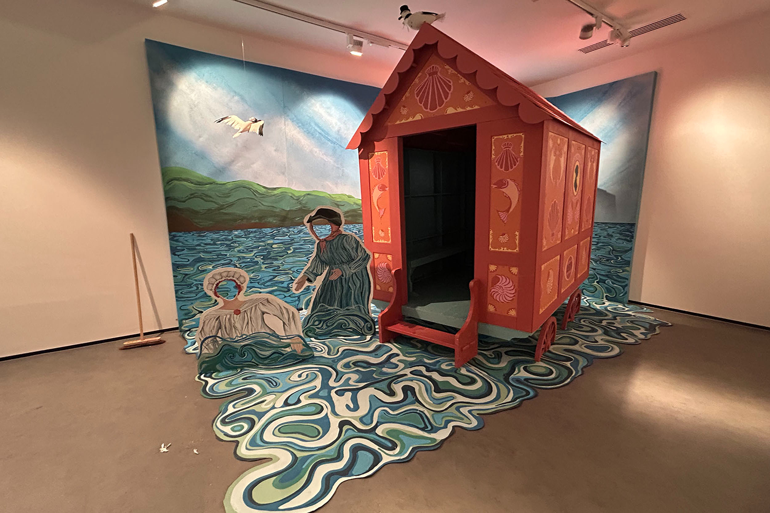 Red wooden caravan positioned in gallery space with painted seascape mural backdrop and swirling blue-green floor pattern beneath.