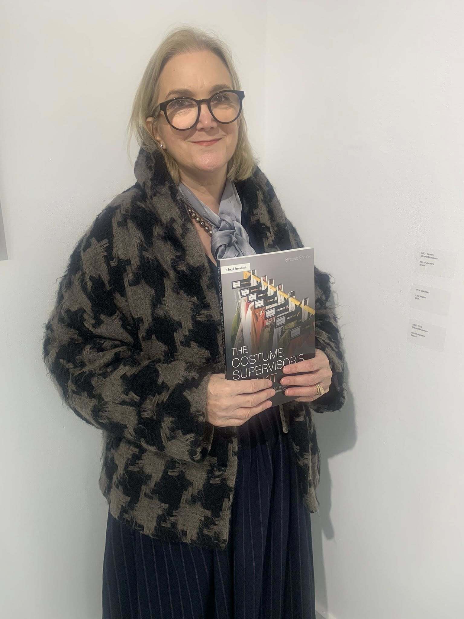 Associate Professor R Pride holding copy of "The Costume Supervisor's Toolkit" book, standing against white wall.