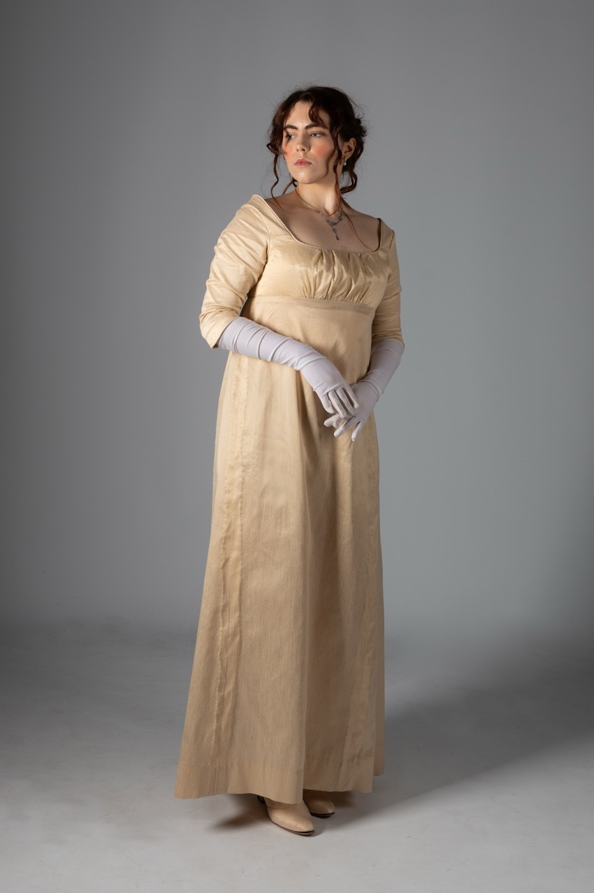 Woman in cream Regency-era gown with empire waist and long white gloves, standing against grey backdrop.