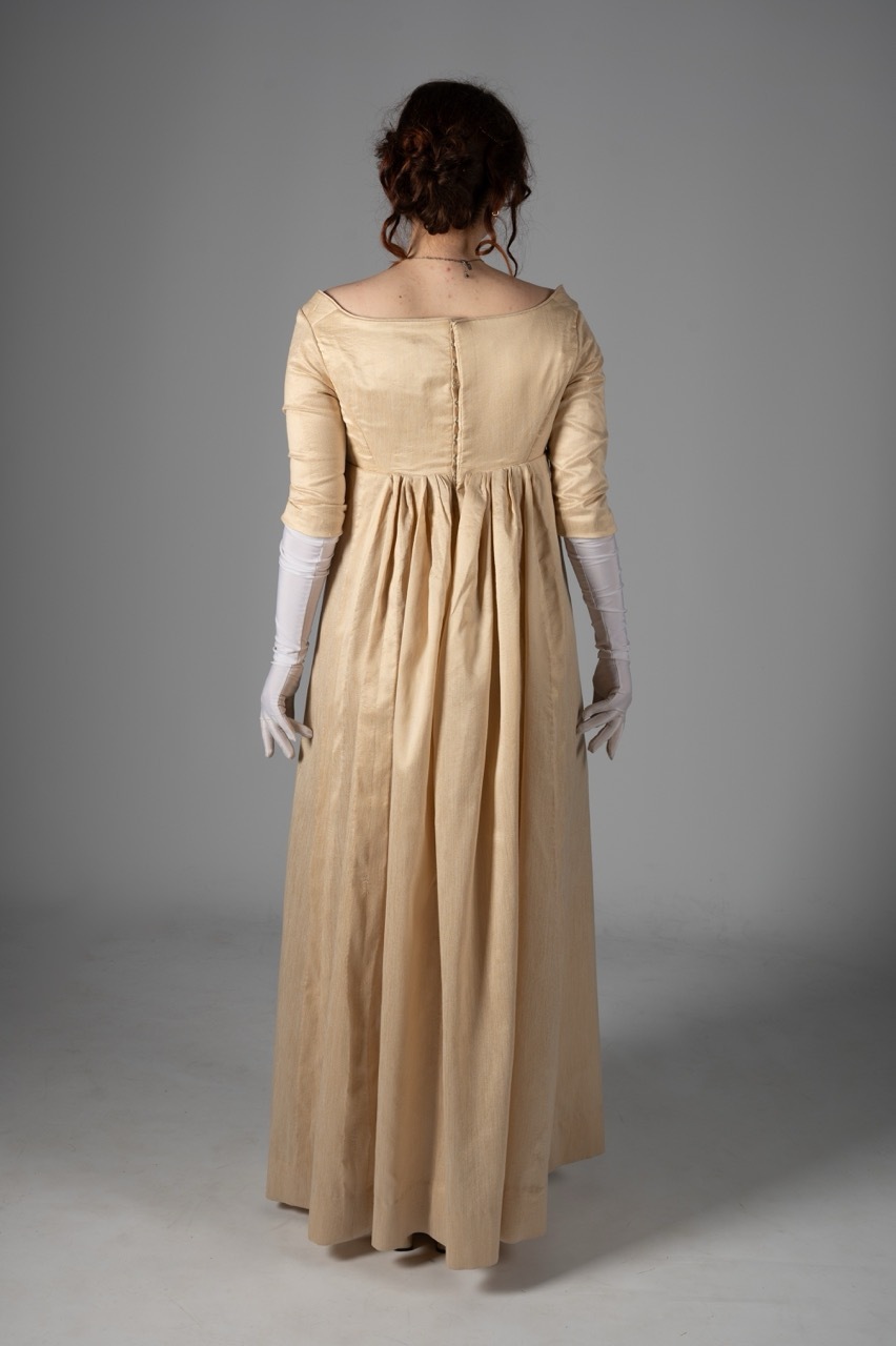 Woman with dark hair in updo wearing cream-coloured empire waist gown and white gloves, shown from behind against grey background.