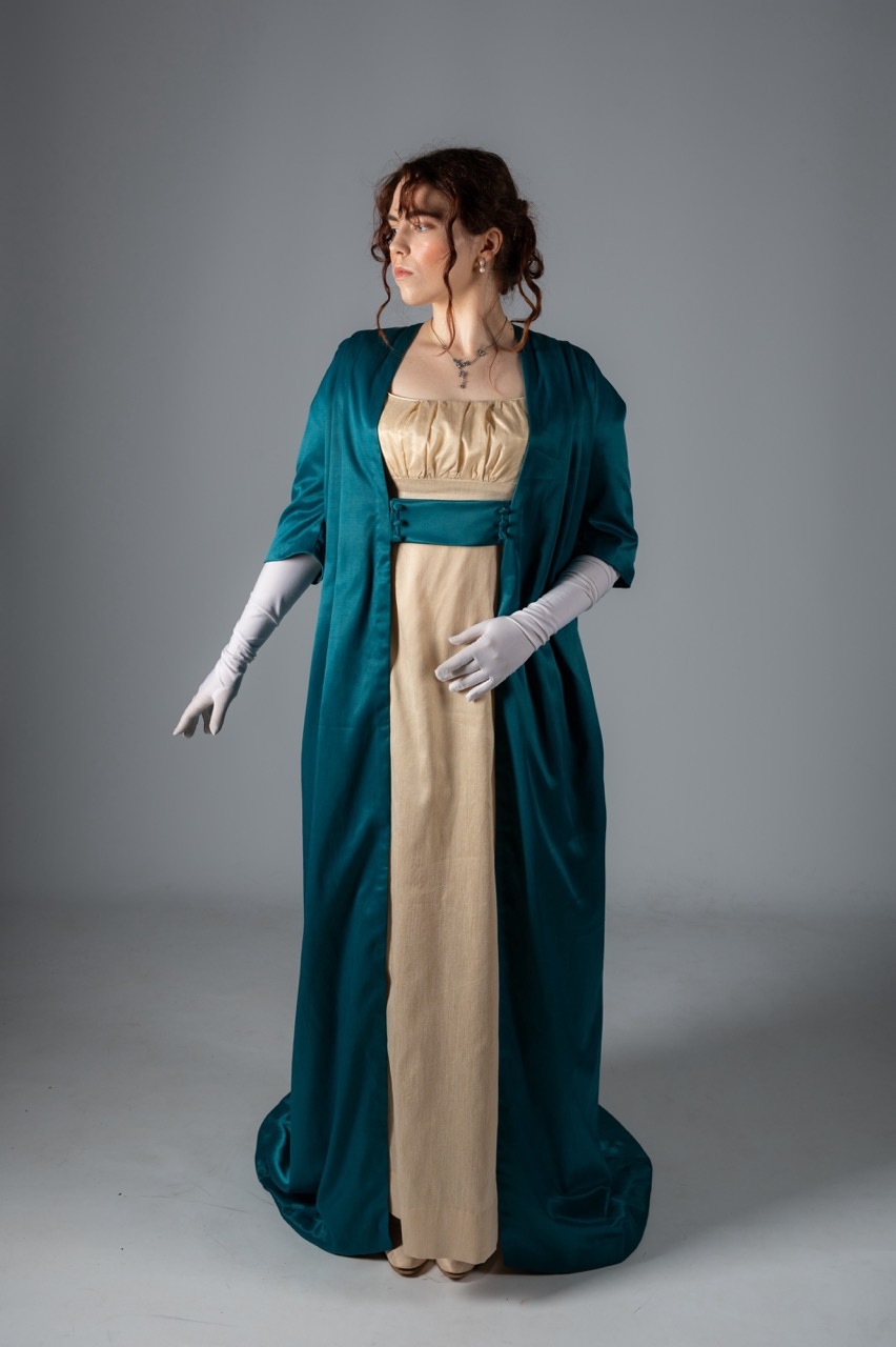 Woman in Regency-era costume: cream empire-waist gown with teal blue spencer jacket and long white gloves, against grey backdrop.