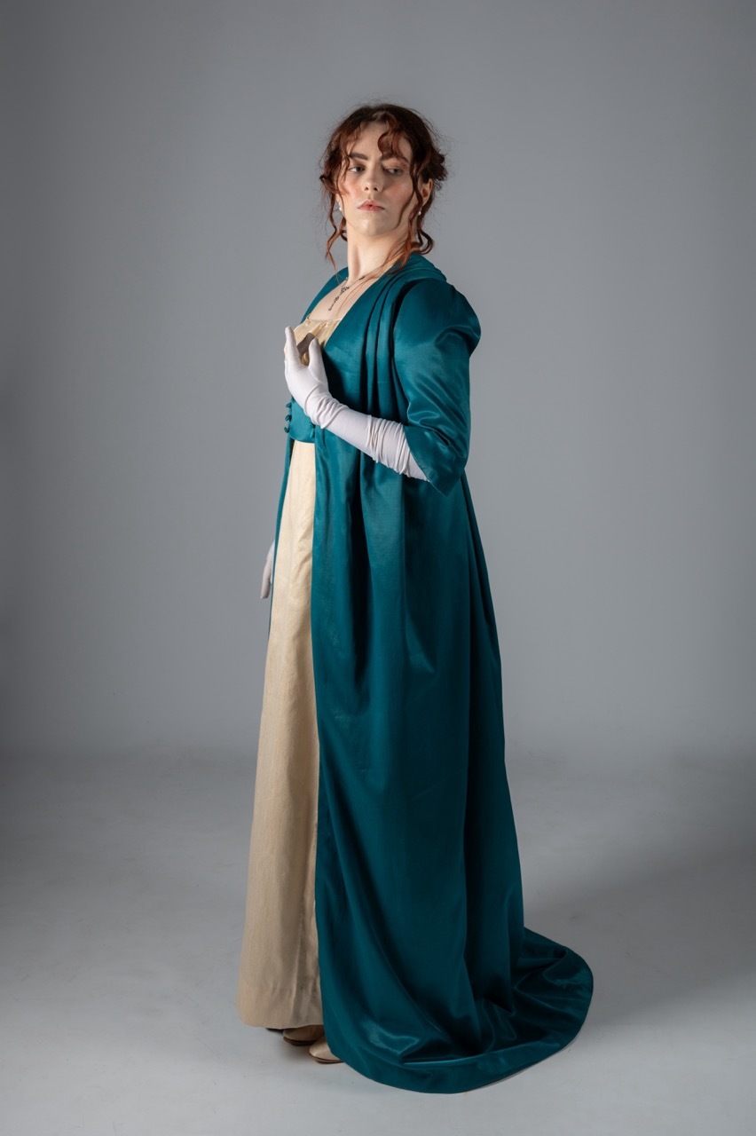 Woman in teal Empire-waist gown with cream underdress and white gloves, standing against grey backdrop in period costume.