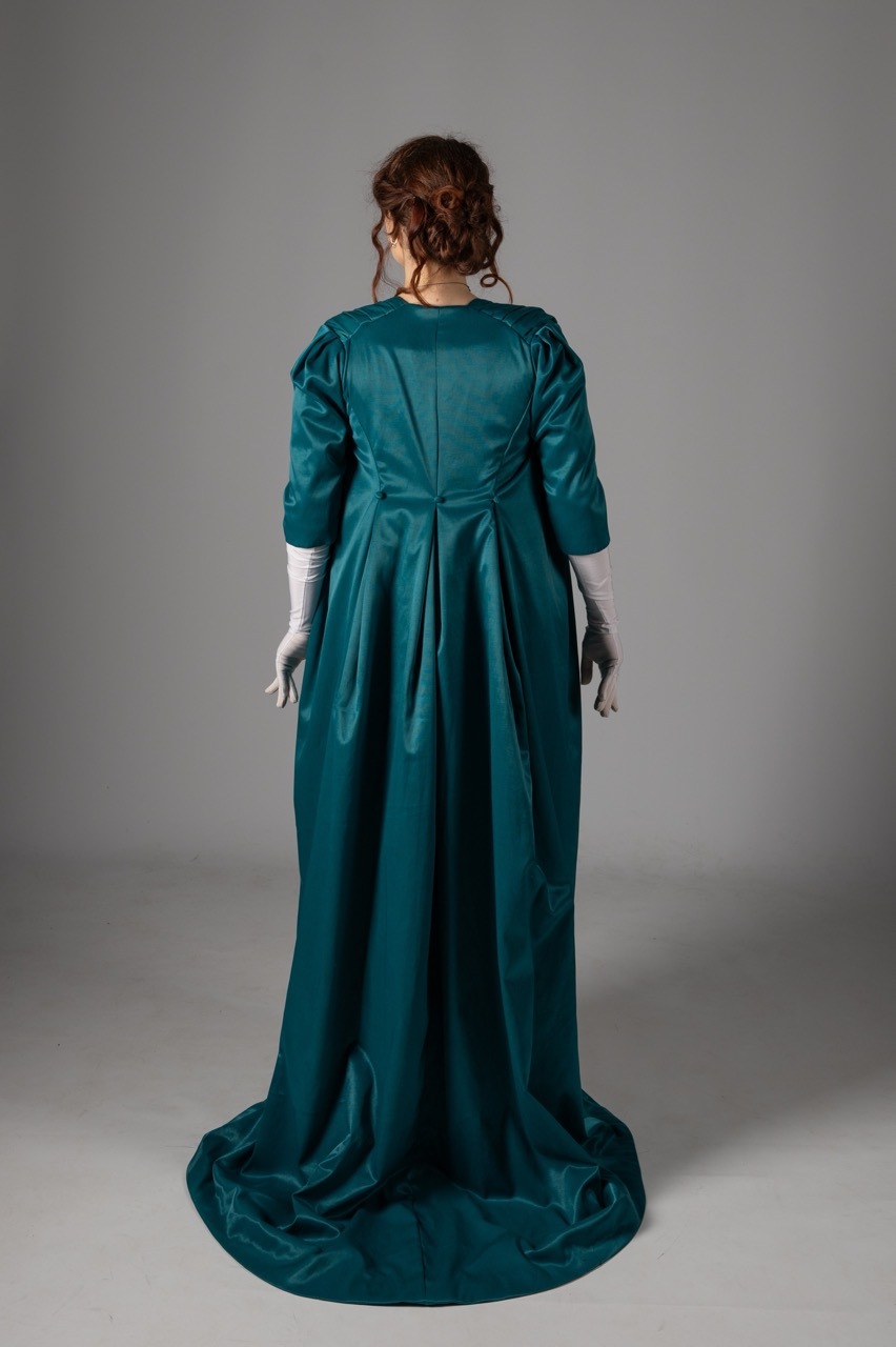 Woman in emerald green silk evening gown with three-quarter sleeves and train, viewed from behind against grey background.