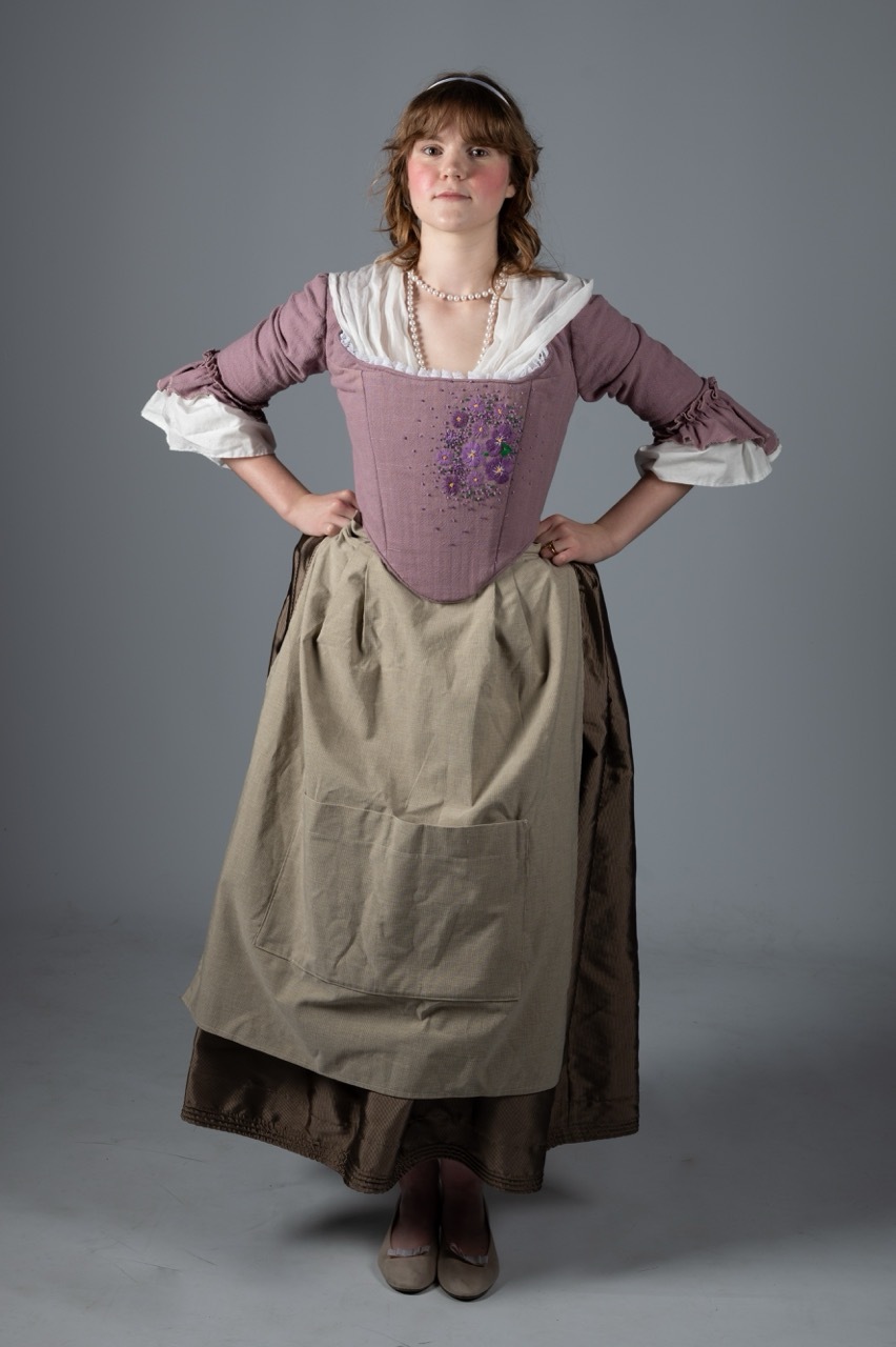 Woman in traditional dirndl with purple bodice, white blouse, and olive green skirt and apron, hands on hips against grey backdrop.