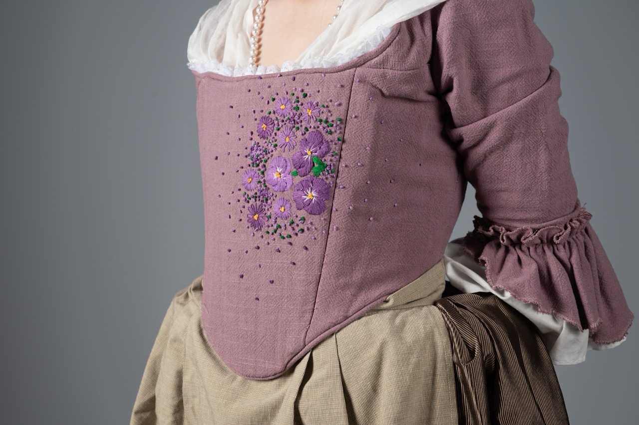 Historical costume with purple bodice featuring scattered beadwork, white chemise, and cream-coloured skirt against grey background.