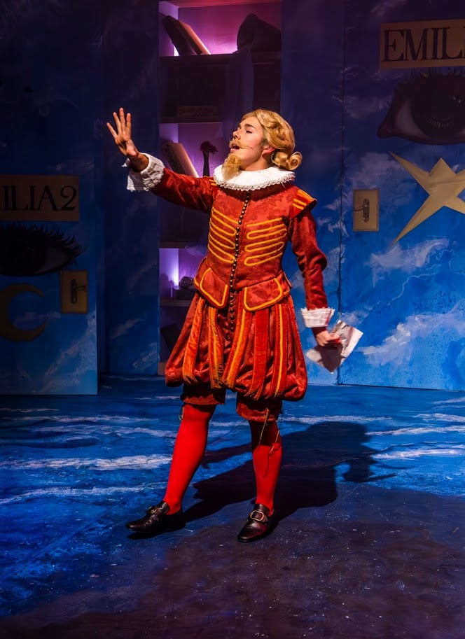 Performer in red Renaissance costume with white ruff collar gesturing on blue stage set with geometric backdrop elements.
