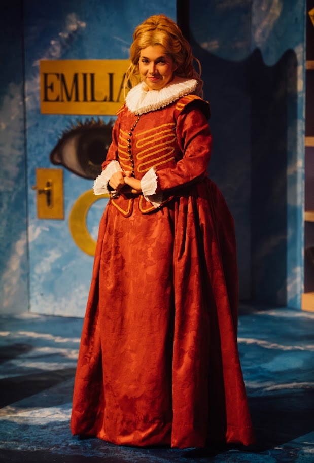 Woman in red Elizabethan-style gown with white ruff collar on stage, yellow "EMILIA" sign visible in background.