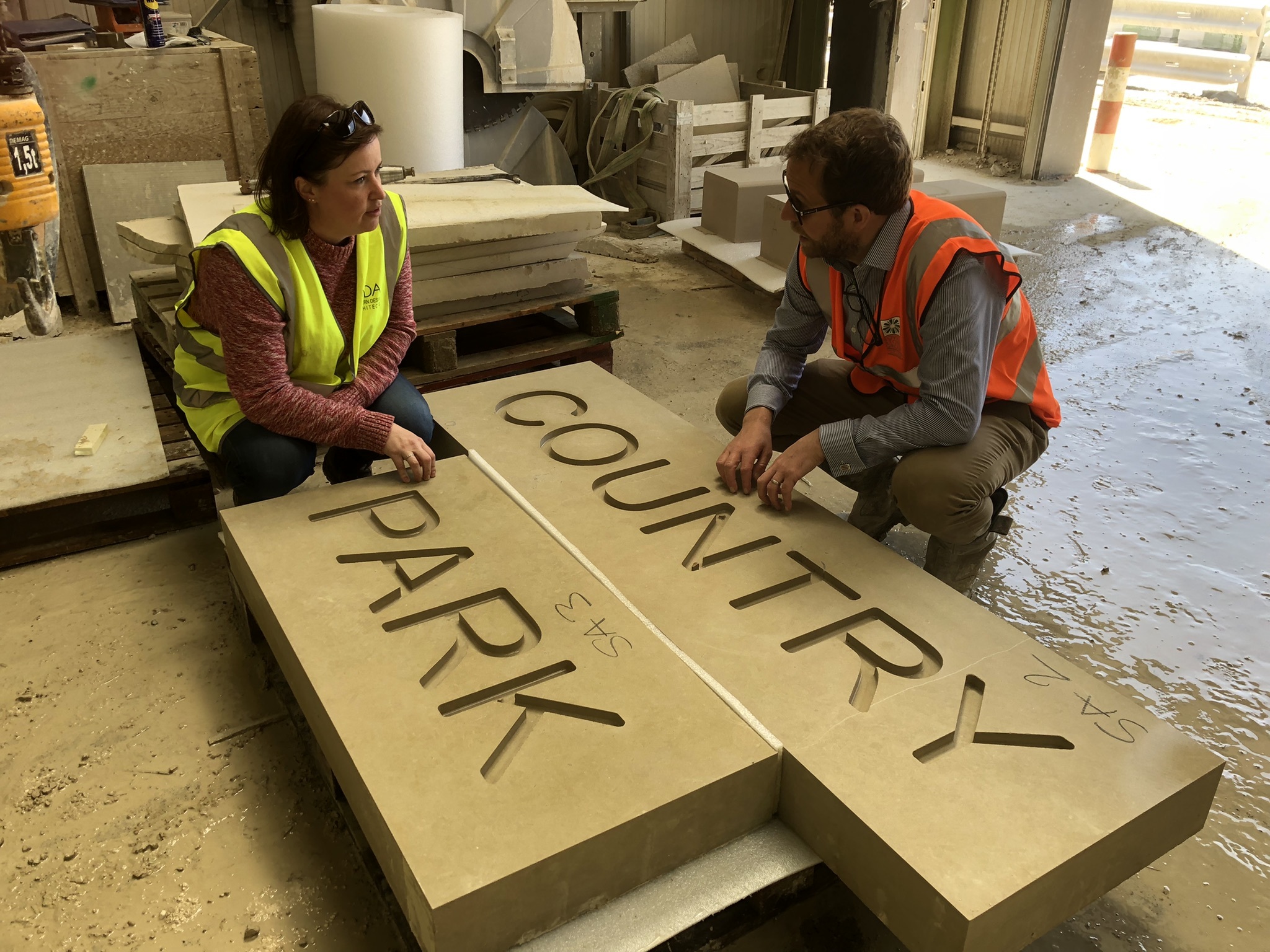 Two designers in hi-viz jackets crouched by a pair of wooden blocks with the words "COUNTRY" and "PARK" engraved on them.
