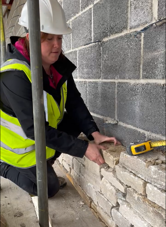 A person wearing a hard hat and hi-viz jacket handling stone bricks as part of a wall.