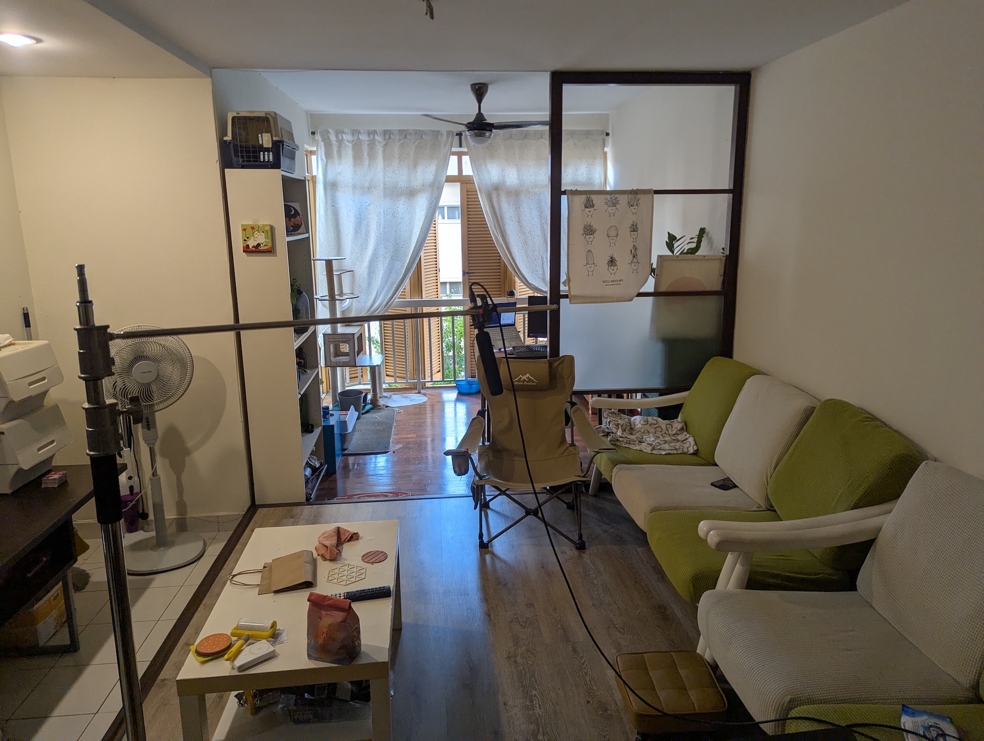 A compact living room with a white and green sofa, a coffee table and a standing fan, opening onto a balcony through sheer curtains.