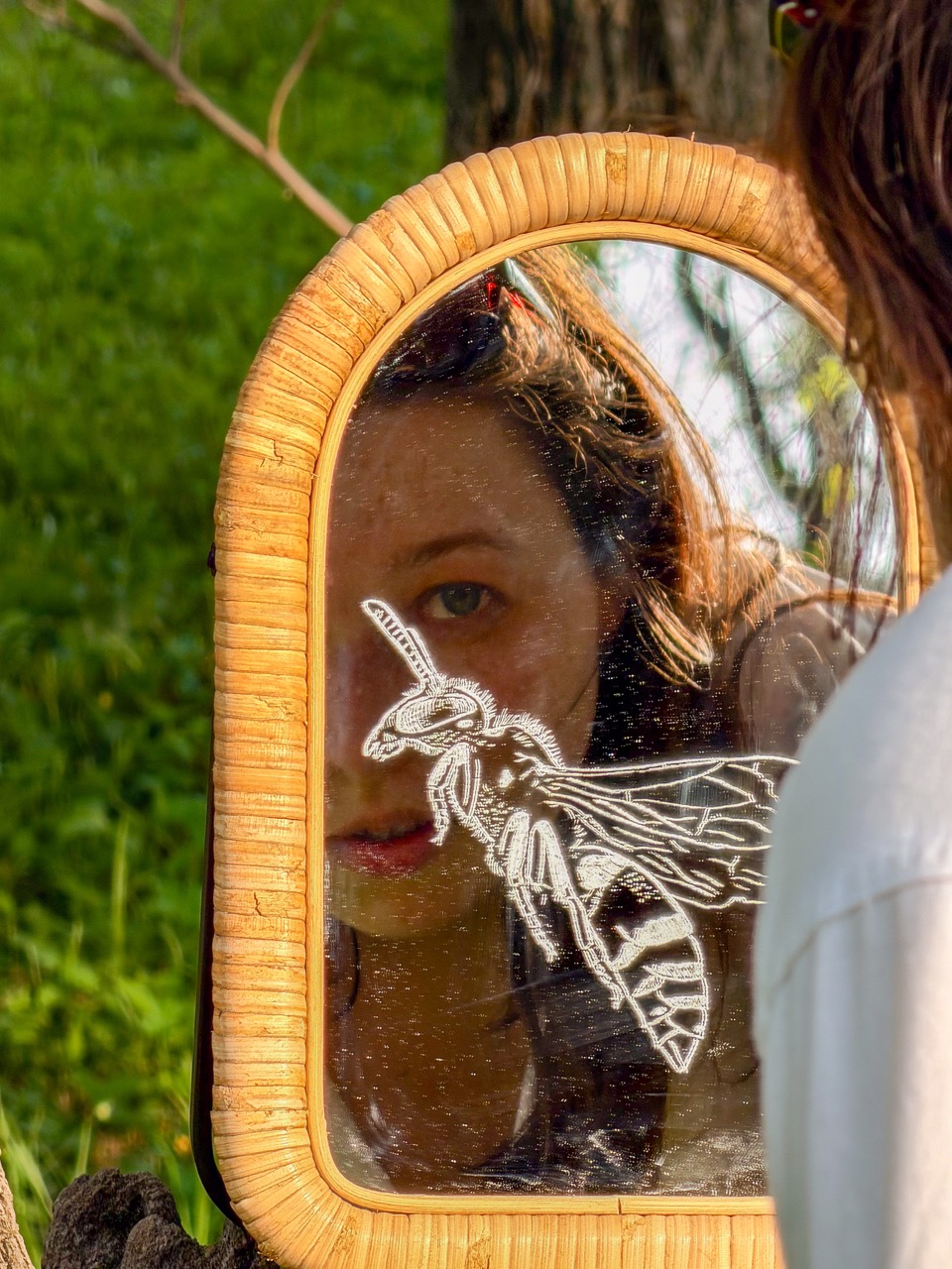 A woman’s face is reflected in a wicker-framed mirror decorated with a wasp illustration, with green grass and a tree trunk in the background.
