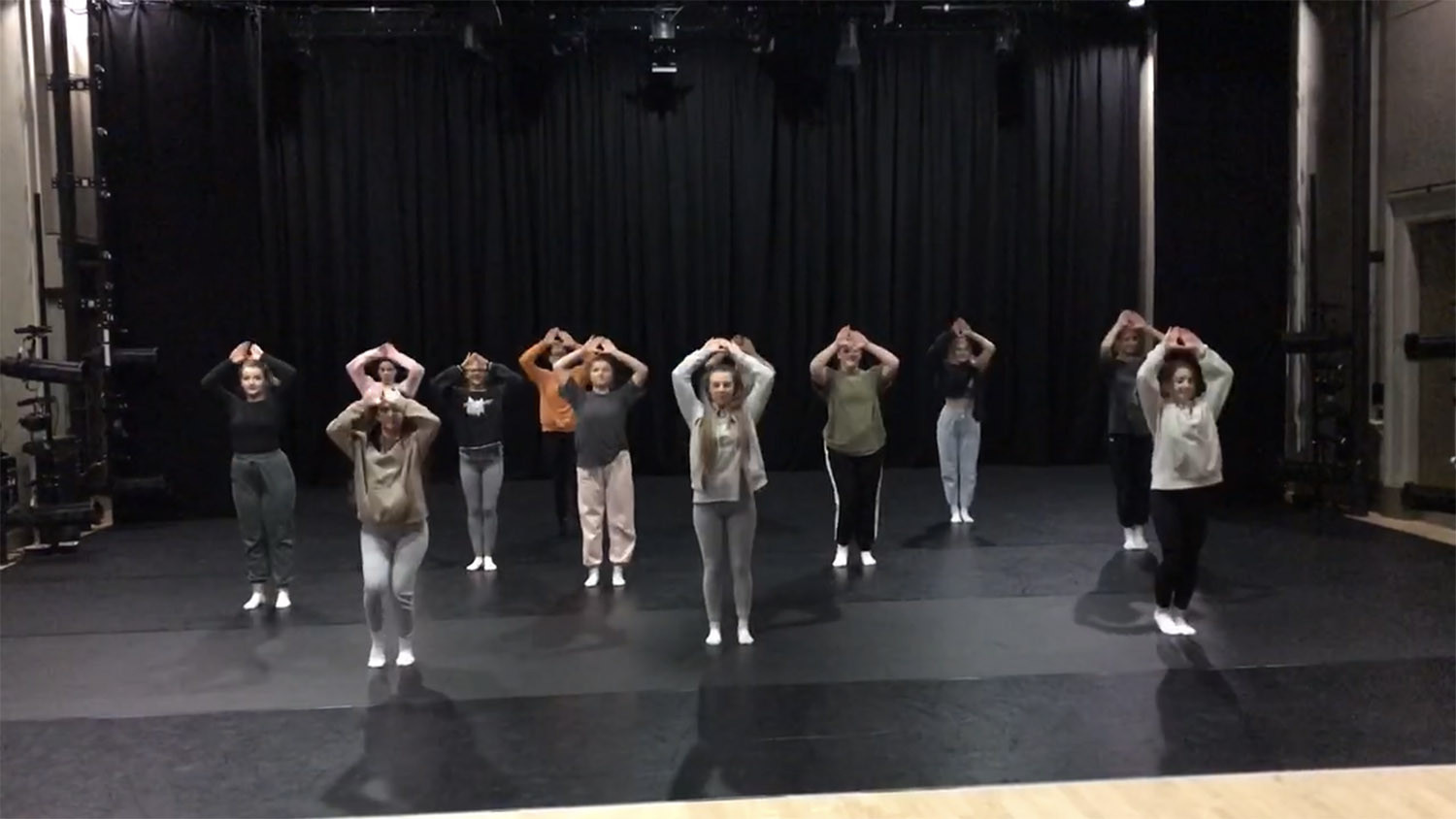 12 dancers rehearsing a performance in a dance studio. The dancers all have their arms raised above their heads.