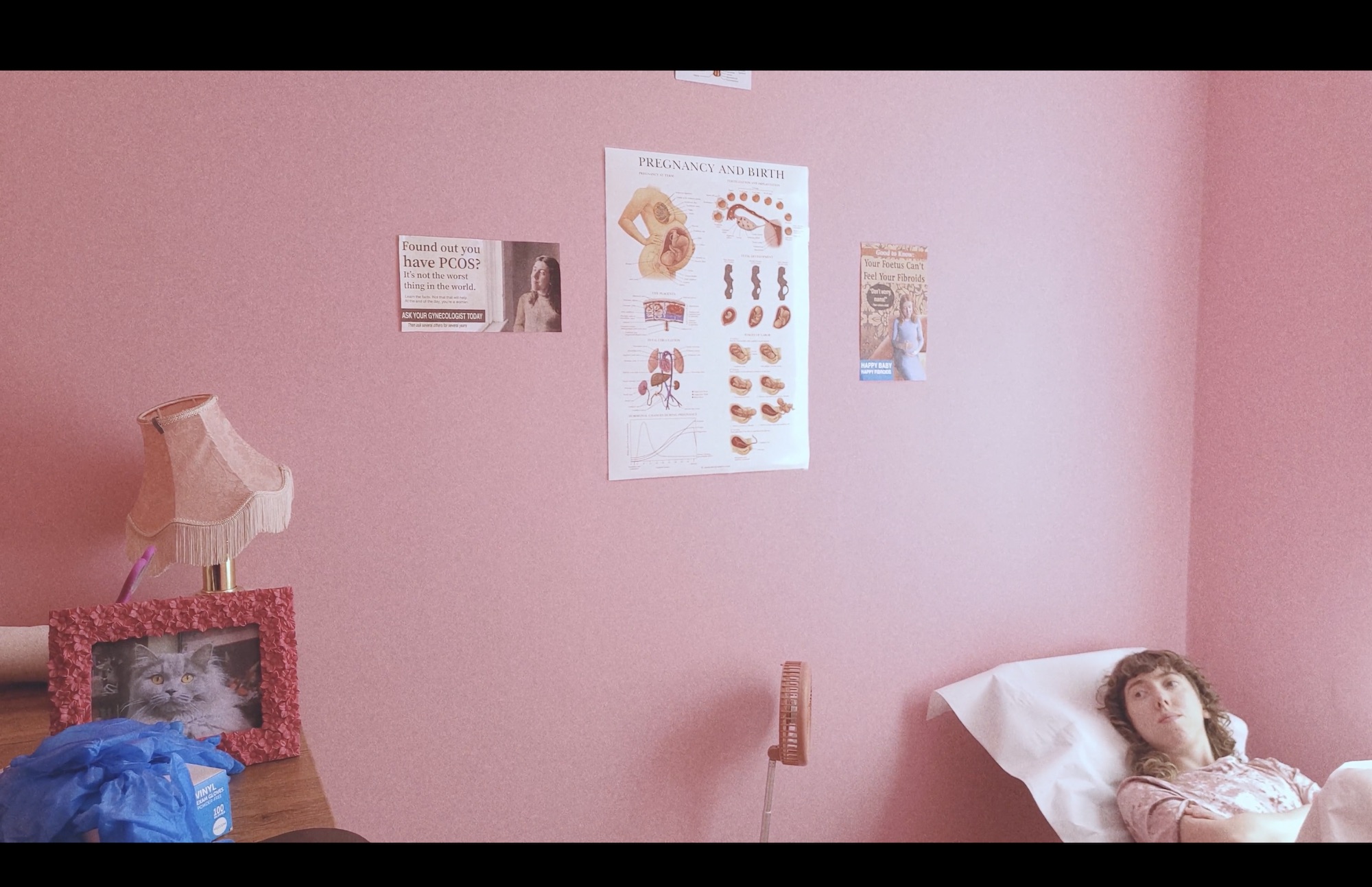 A woman lies on a white bed beside a pink wall with posters and a bedside table holding a lamp and framed photo.