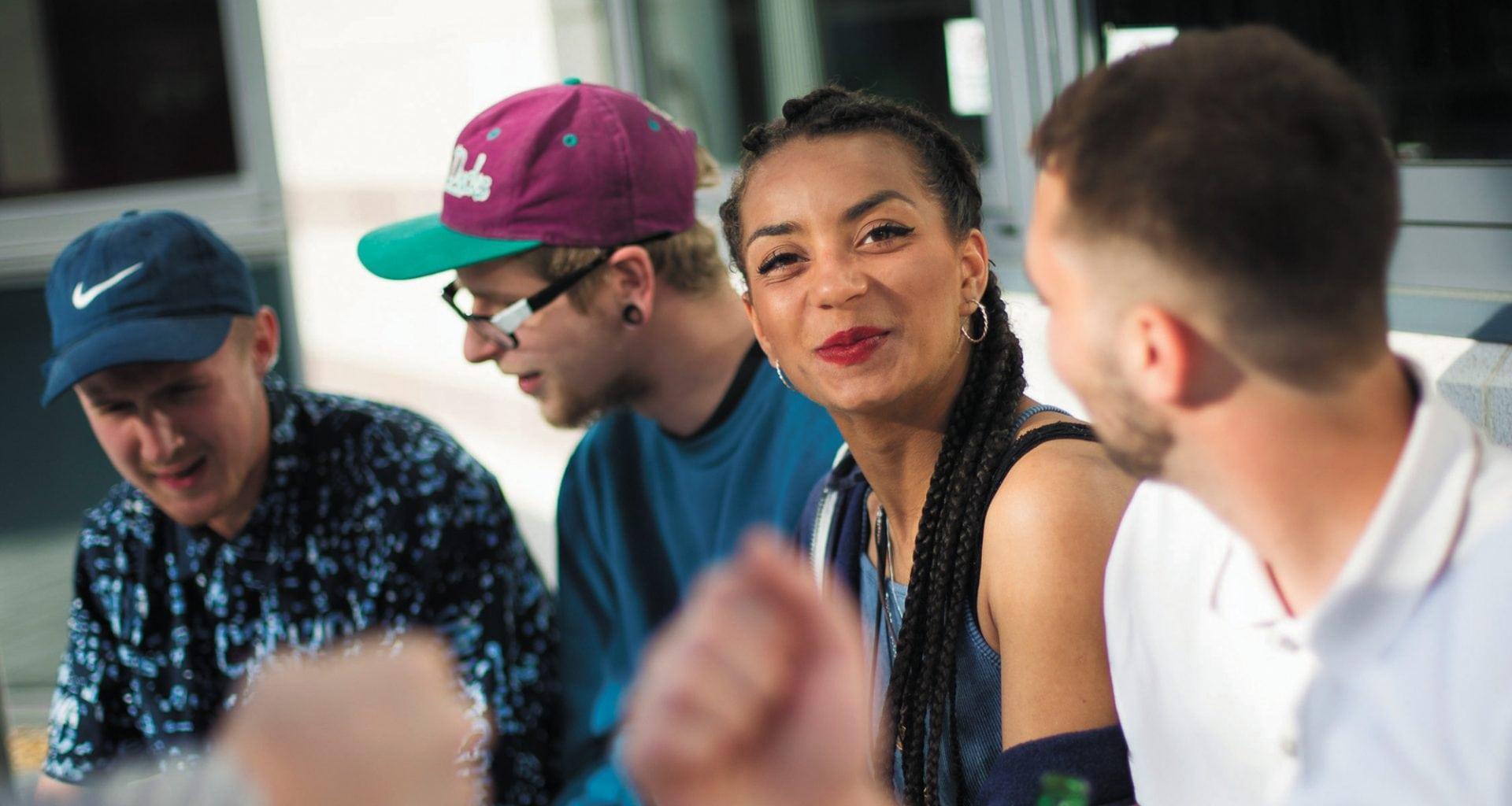 Four students sitting and talking, a woman in the group is looking to the camera and smiling