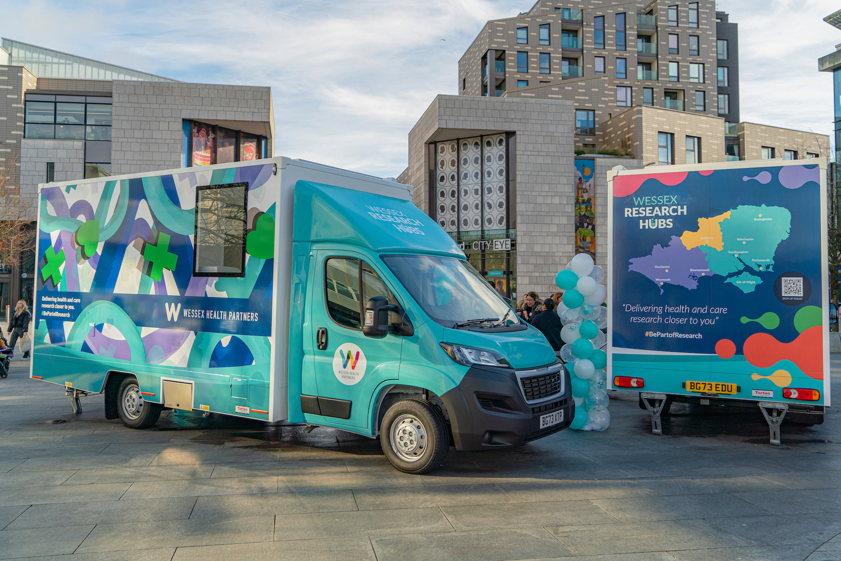 Two NHS/NIHR research buses on display in Southampton's Guildhall Square. One of the buses is viewed from the back, showing a colourful map of the Wessex area.