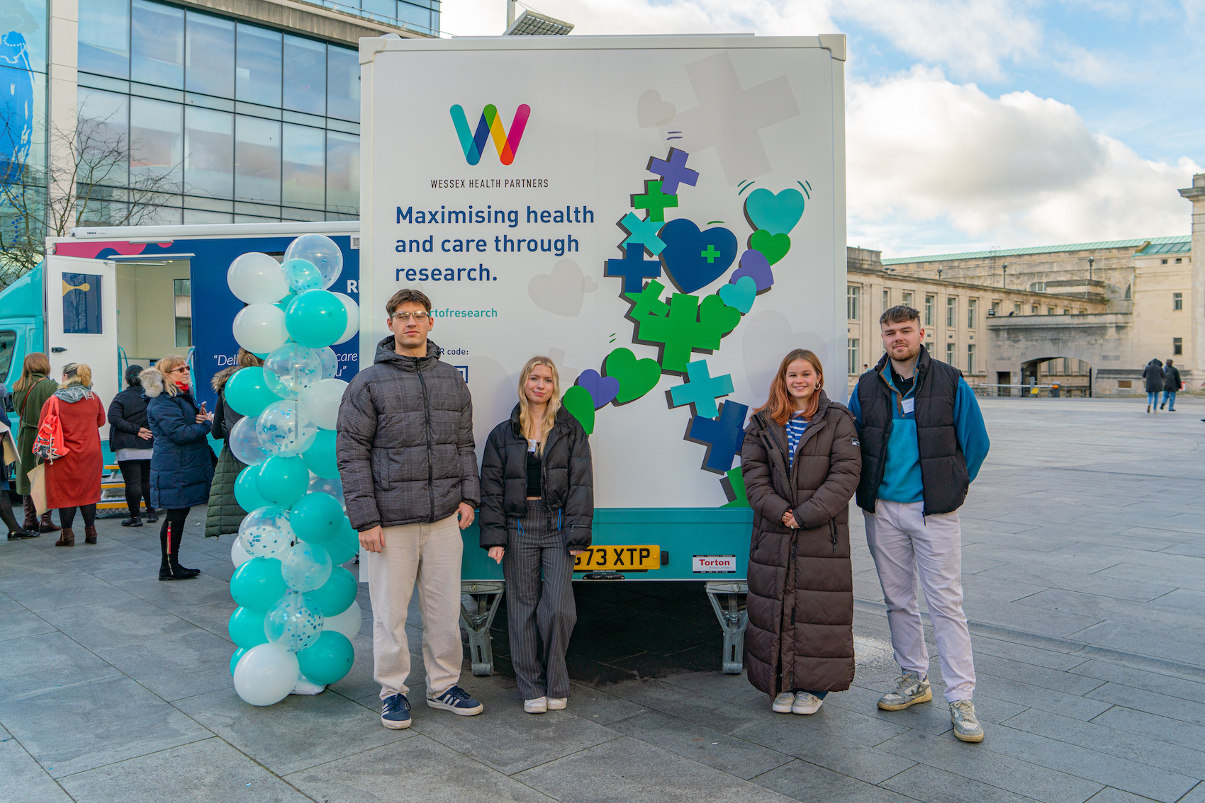 Four BA (Hons) Interior Architecture and Design students pose for a photo in front of an NHS/NIHR Research Bus in Southampton. The bus displays the mascot "Maxwell".