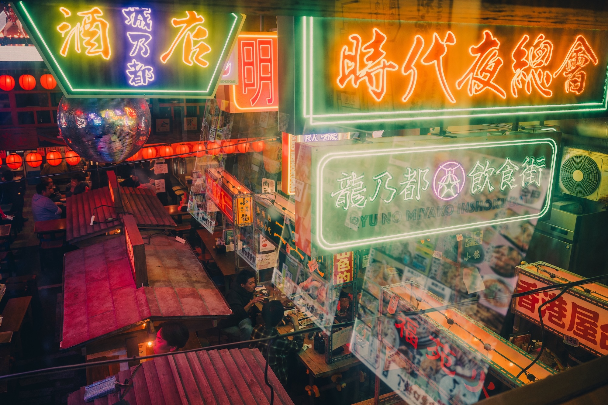 A vibrant urban scene featuring illuminated neon signs in different colours. The signs advertise various eateries and establishments, while tables are arranged underneath. The perspective shows a bustling atmosphere filled with lanterns and seating areas.