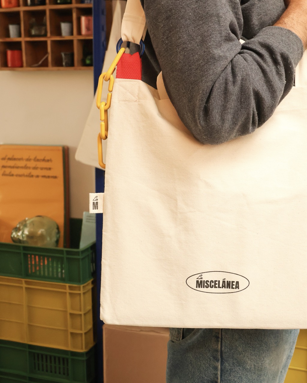 A person carrying a cream Miscelánea tote bag with yellow handles, standing beside stacked plastic crates and shelves in a shop.