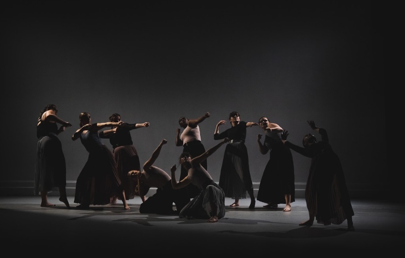 A large group of dancers in a dimly lit stage. They are wearing long skirts and dark Lacy tops. Some are standing, some are kneeling. Each dancer has one arm pulled back close to their head, and their other arm is extended forward, as in the action of drawing a bow.