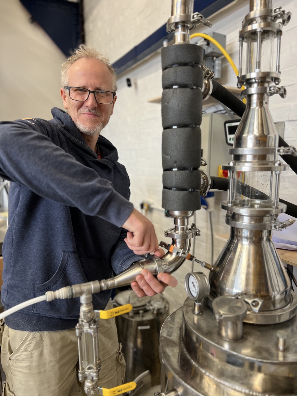 Man smiling, stood by silver distillery equipment in a white room.
