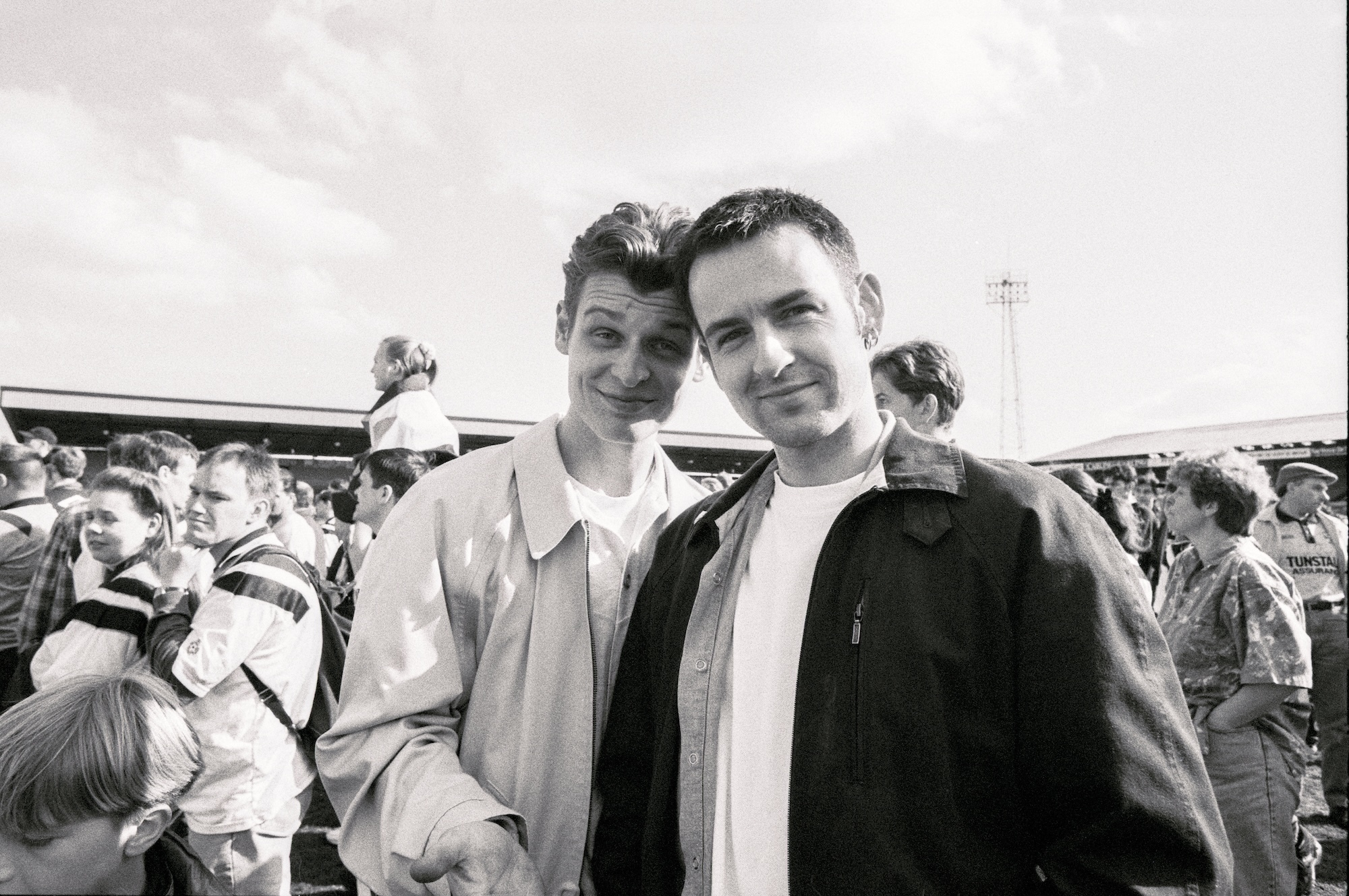 Black and white photo of two men posing to the camera while at a football match.