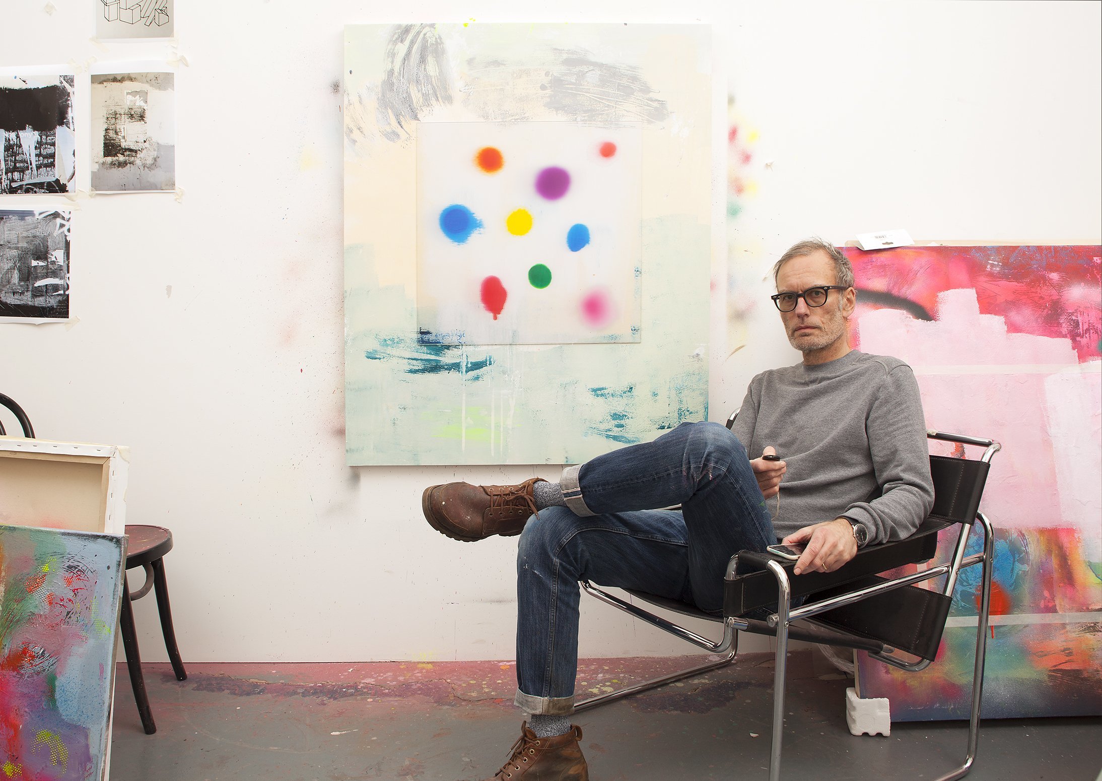 R Paul sitting in a gallery space with canvases behind him in the background.