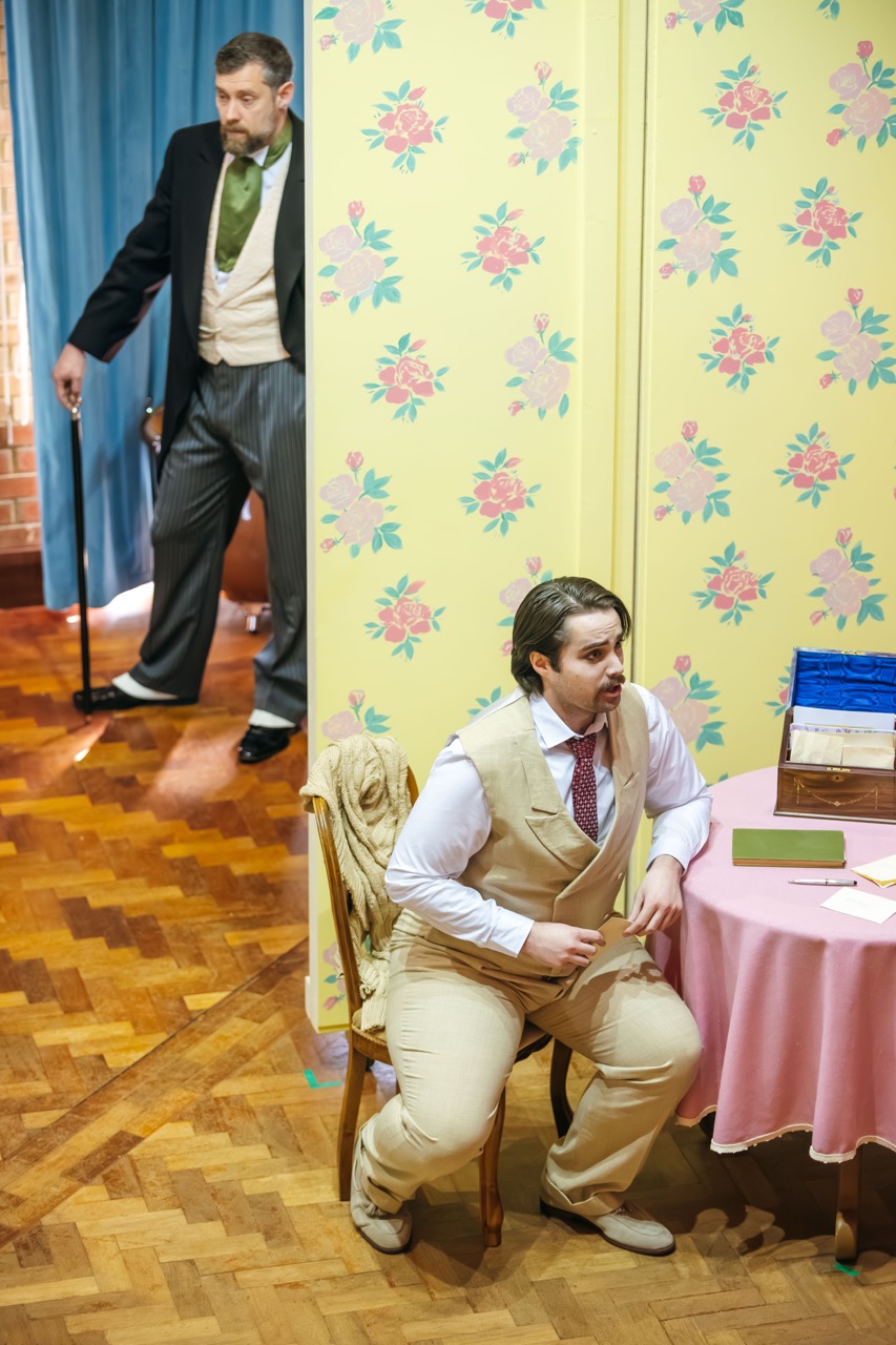 A man in formal attire stands behind a floral-patterned wall, while another man in a beige suit sits on a chair at a small table covered with a pink tablecloth, holding a piece of paper. The setting features wooden flooring with warm tones.