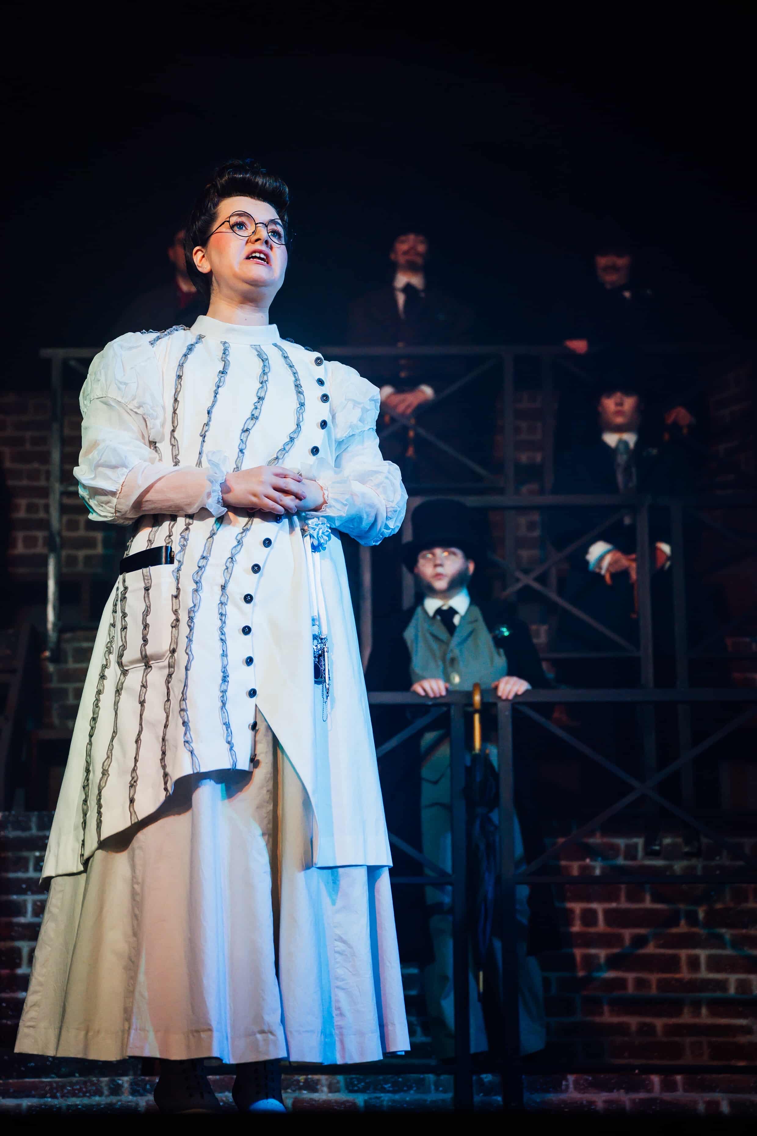 A performer in a vintage white dress with black buttons stands centre stage, appearing to deliver lines. In the background, three men in dark suits observe from a balcony-like structure, partially obscured in shadow.