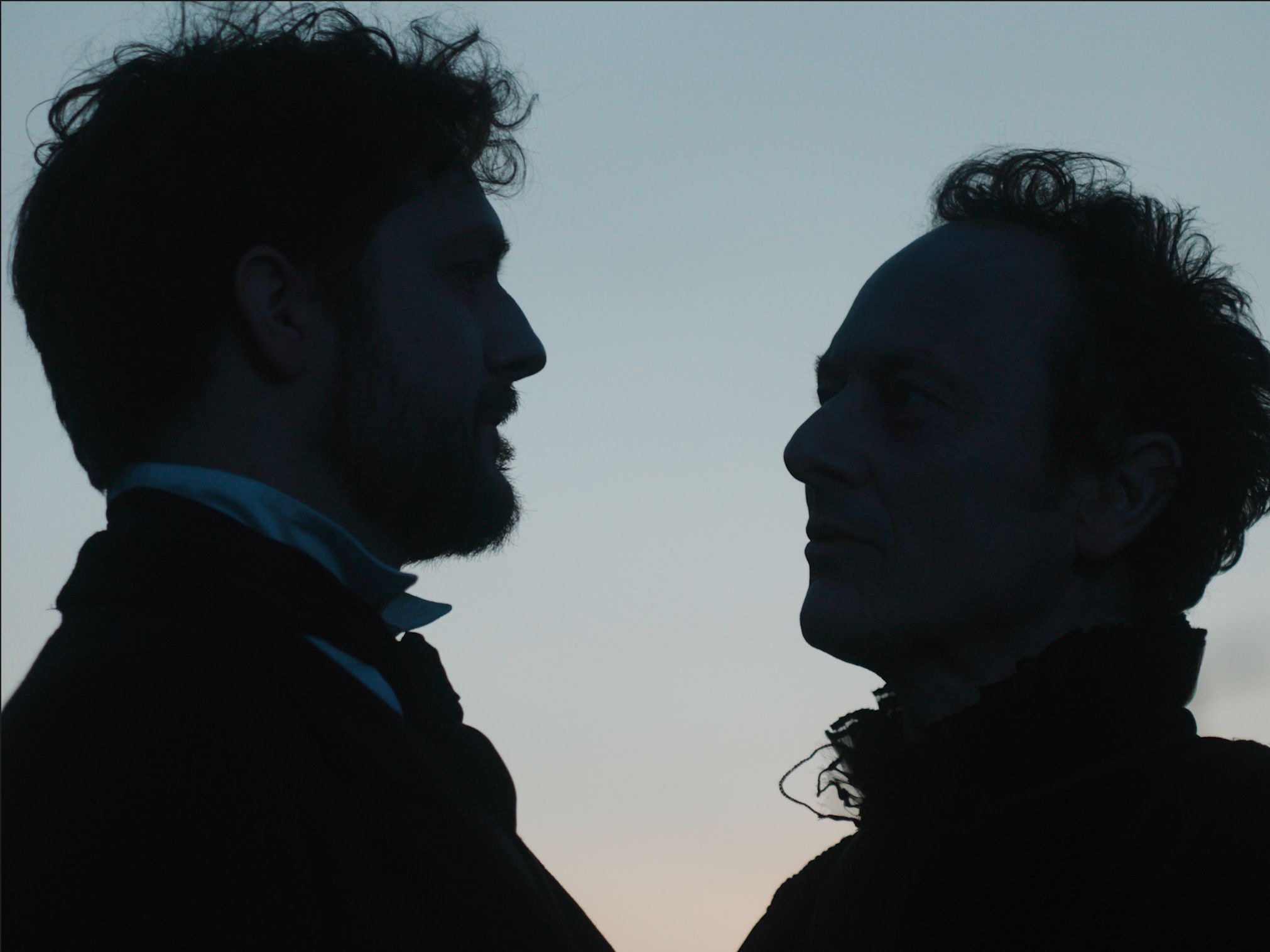 Silhouettes of two men standing face to face against a twilight sky. The man on the left has curly hair and is dressed in a dark outfit with a white collar, while the man on the right has a more angular face with tousled hair, both facing each other.