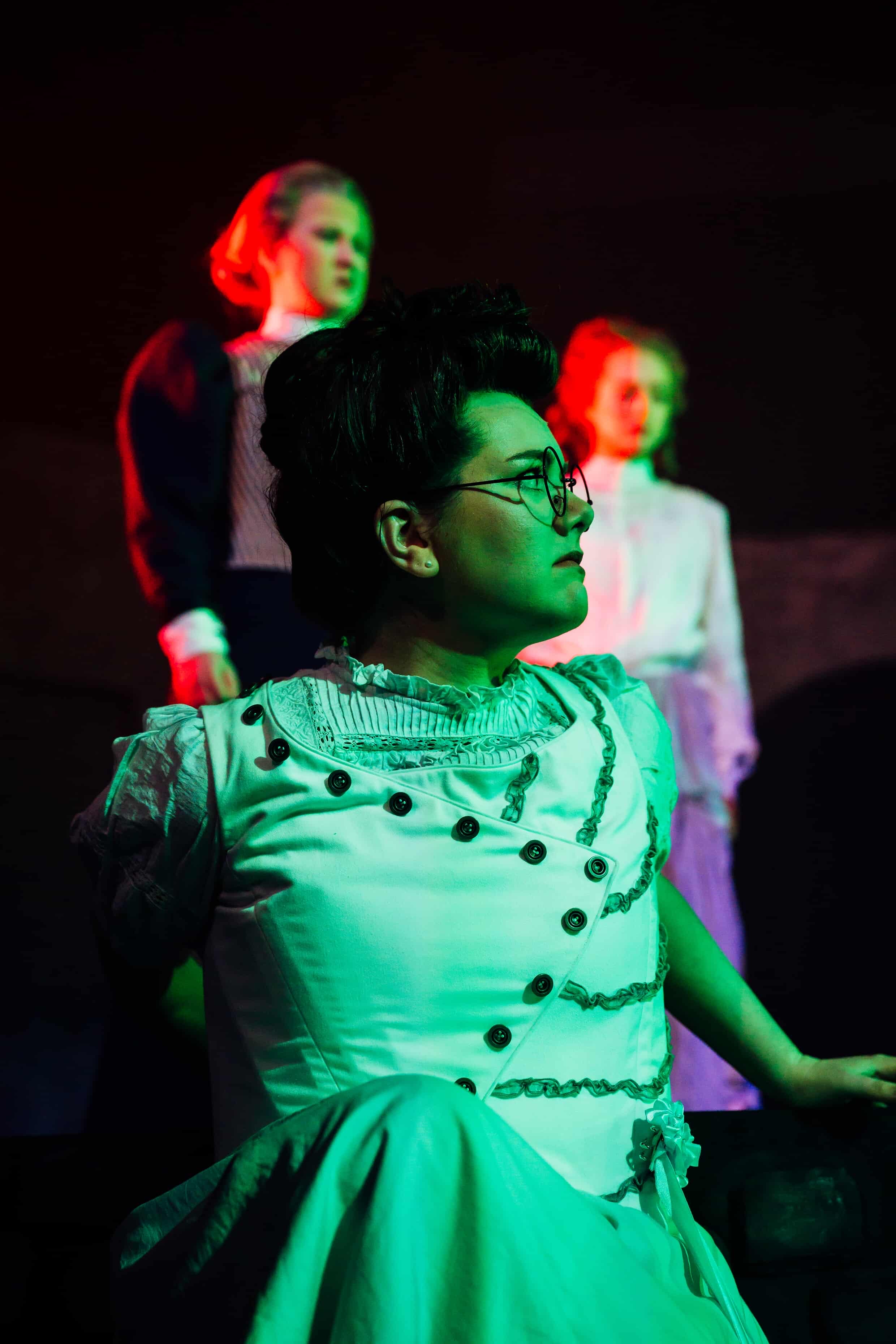 A woman wearing a historical-style white dress and glasses sits in the foreground, while two individuals in period costumes appear blurred in the background. The scene is lit with green and red lighting, creating a dramatic effect.