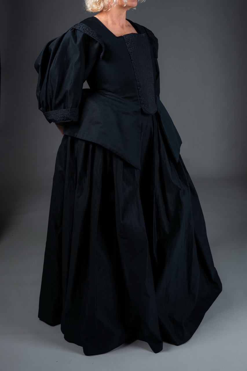 A woman dressed in a historical black gown with puffed sleeves and a flared skirt. The gown features a fitted bodice and is designed in a period style, set against a plain grey background.