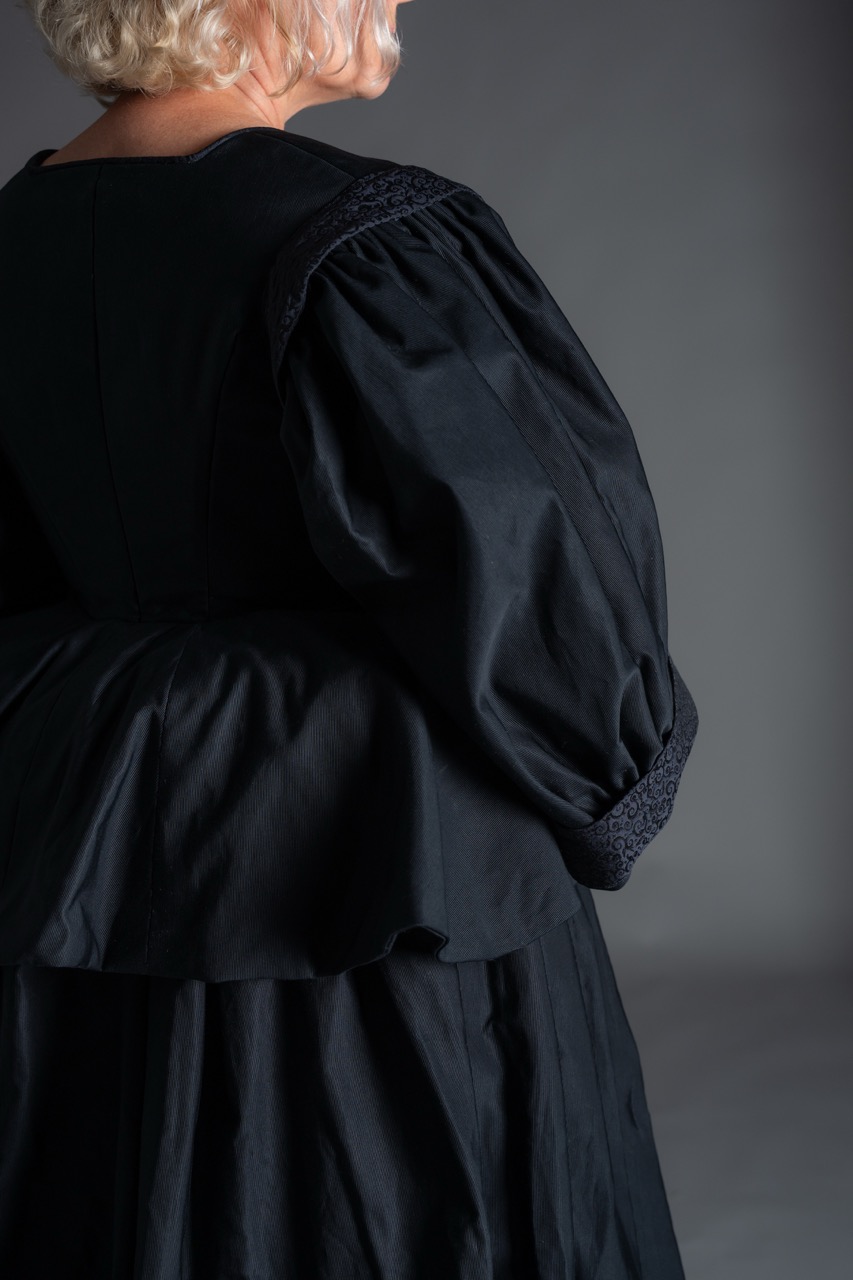 A woman wearing a black dress with a puffed sleeve detail is shown from the side. The dress features a fitted bodice and a flowy peplum style, while the background is a neutral grey.