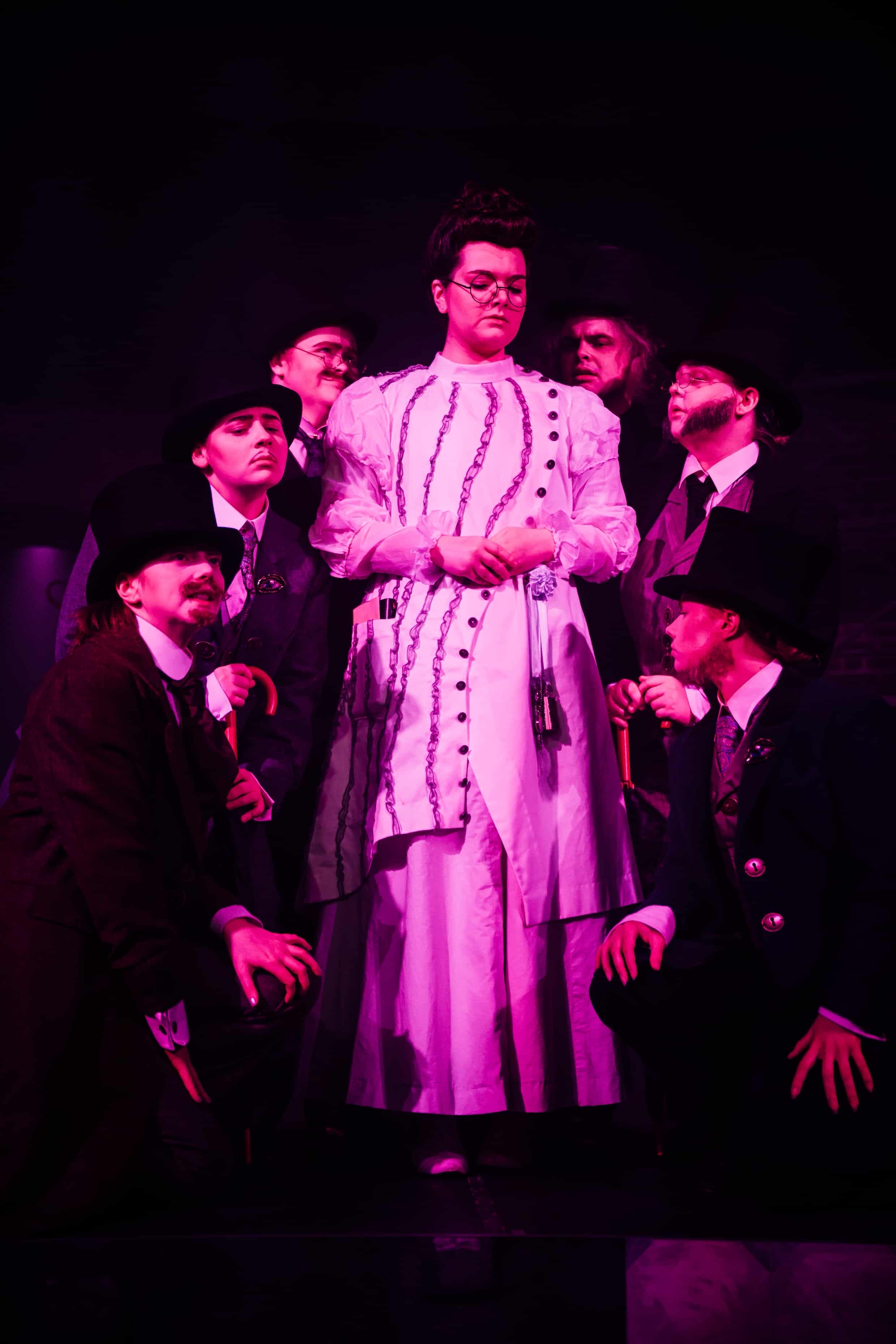 A group of performers is depicted on stage, with a central figure in a long white dress and glasses, surrounded by six men in suits and hats. The lighting is dark, creating a dramatic atmosphere with purple hues.