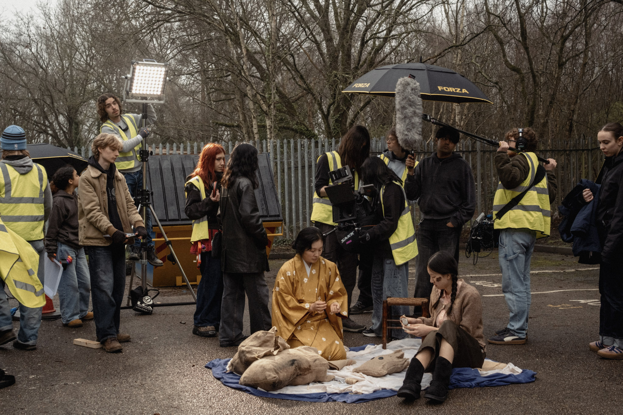 A film set with a group of crew members in high-visibility jackets, working on a shoot. Several people are positioned around a blue blanket on the ground, while some hold microphones and cameras, with trees and a fence in the background.