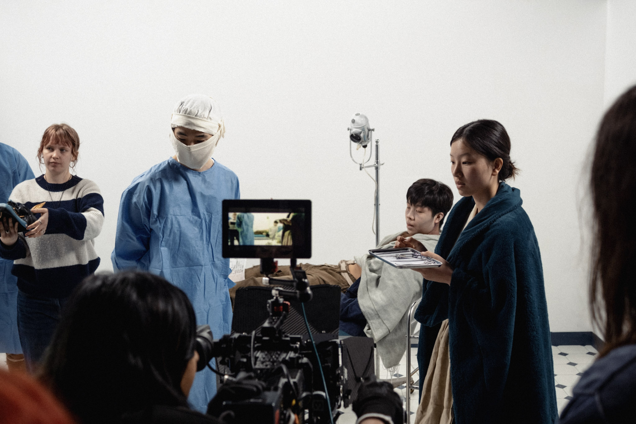 A group of people on a film set, including a woman holding a tablet and wearing a green shawl, a man in a surgical mask and blue gown, and others behind a camera. The background is a plain wall, and there is a light stand nearby.