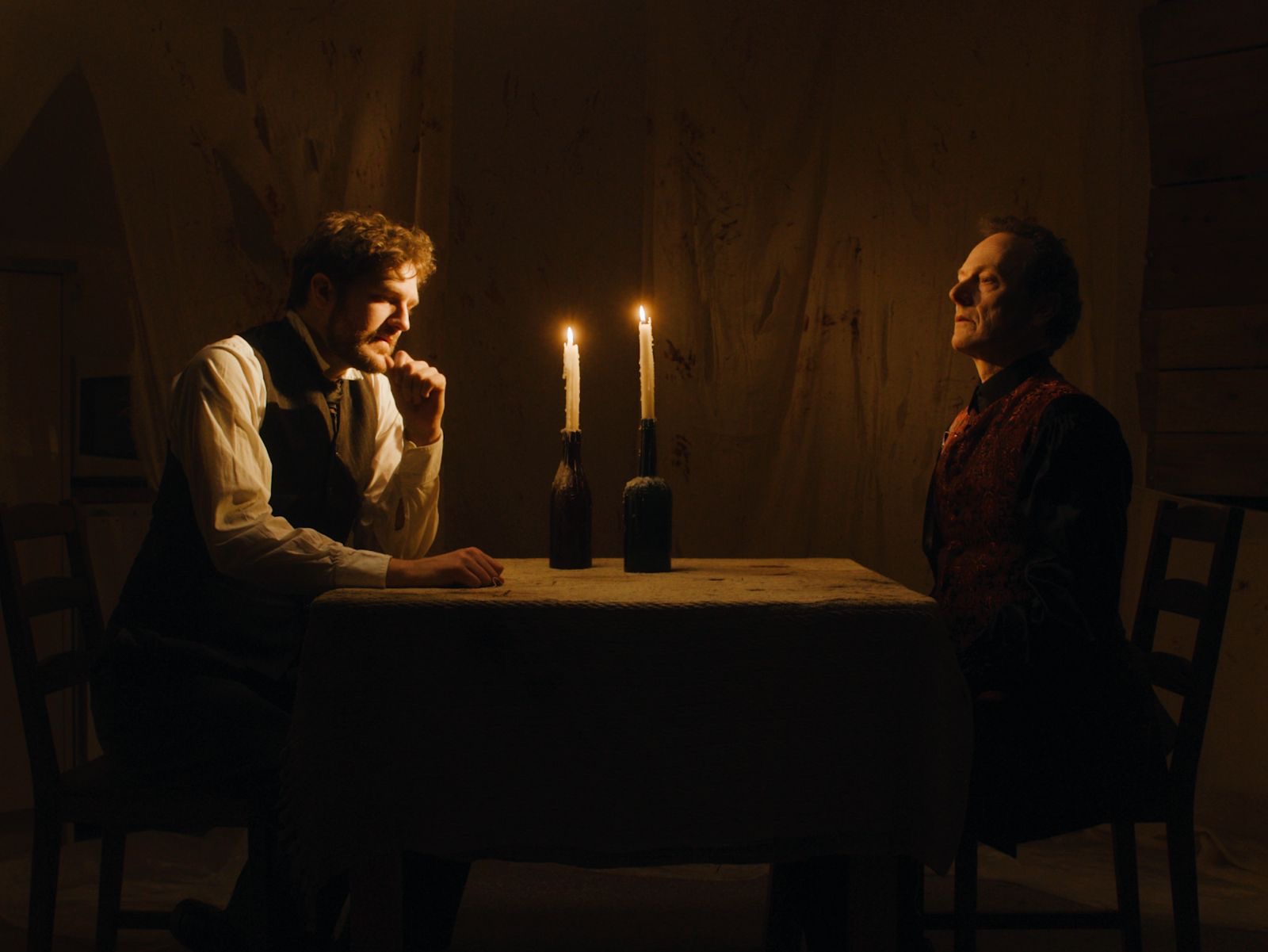 Two men seated at a small table covered with a cloth in a dimly lit room. A lit candle stands on the table between them, while soft light casts shadows on the wall behind, enhancing the intimate atmosphere.