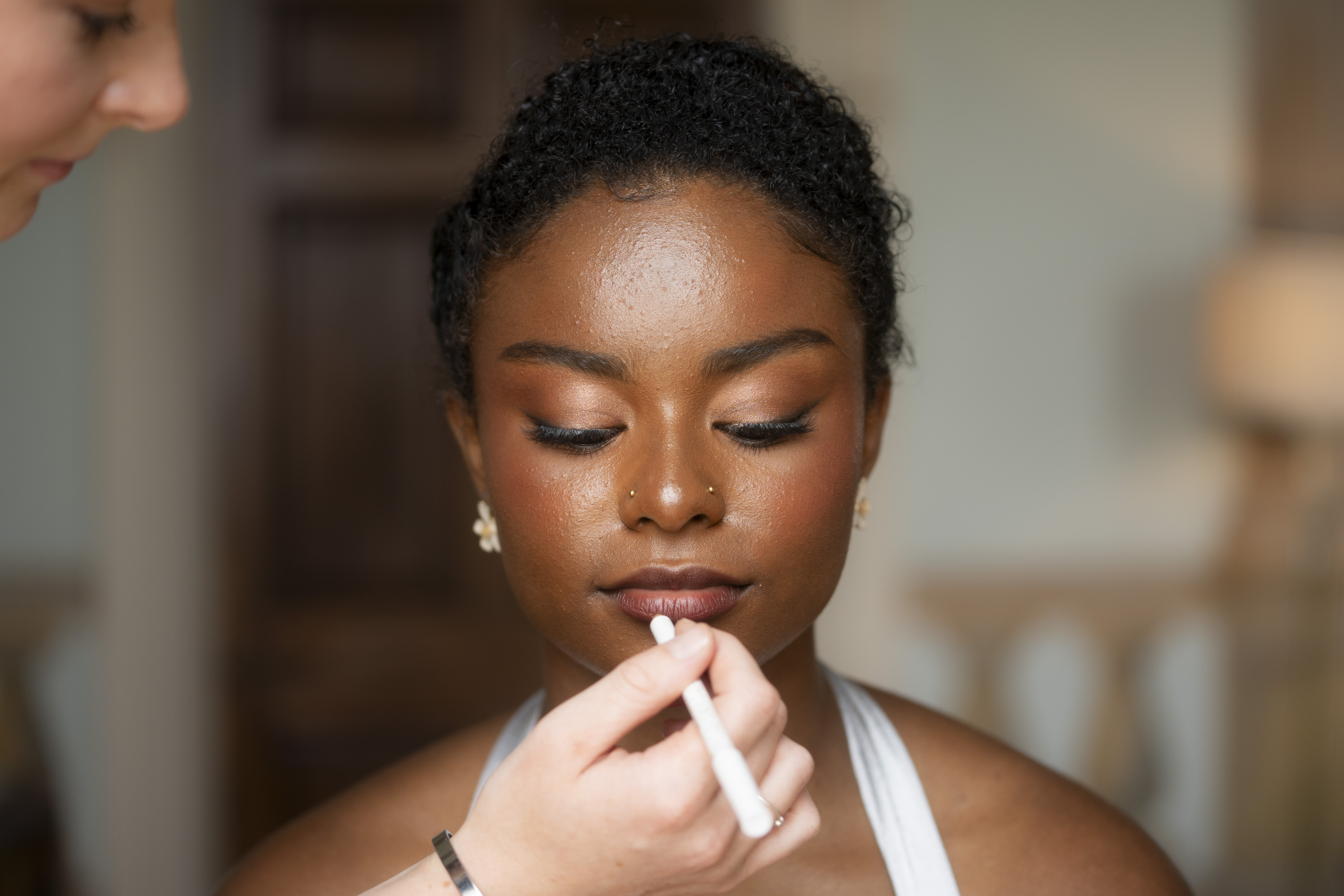 Woman having lipstick applied by makeup artist, eyes closed, wearing pearl earrings in softly lit interior setting.