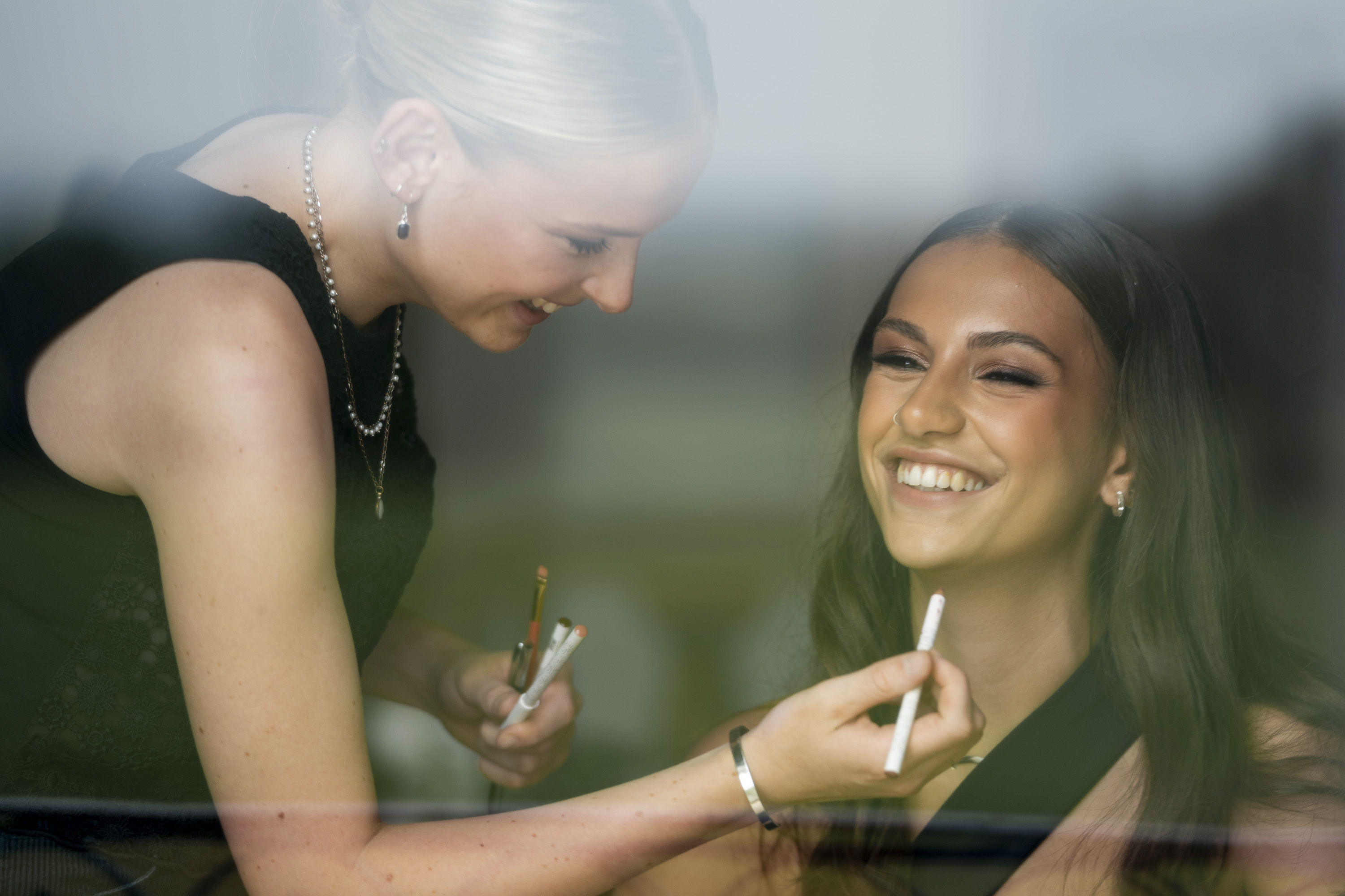 Two women laughing together inside a car, one leaning towards the other, both holding cigarettes or similar items.