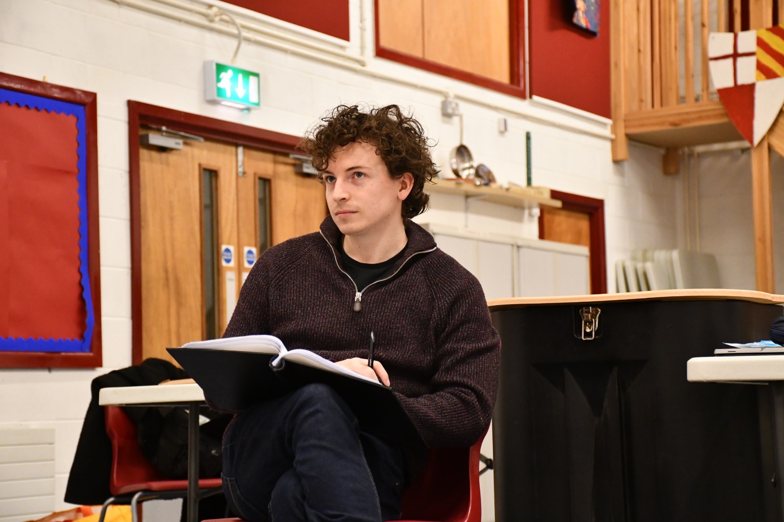Young person with curly hair in dark hoodie sits reading book in modern library with wooden upper level and white walls.