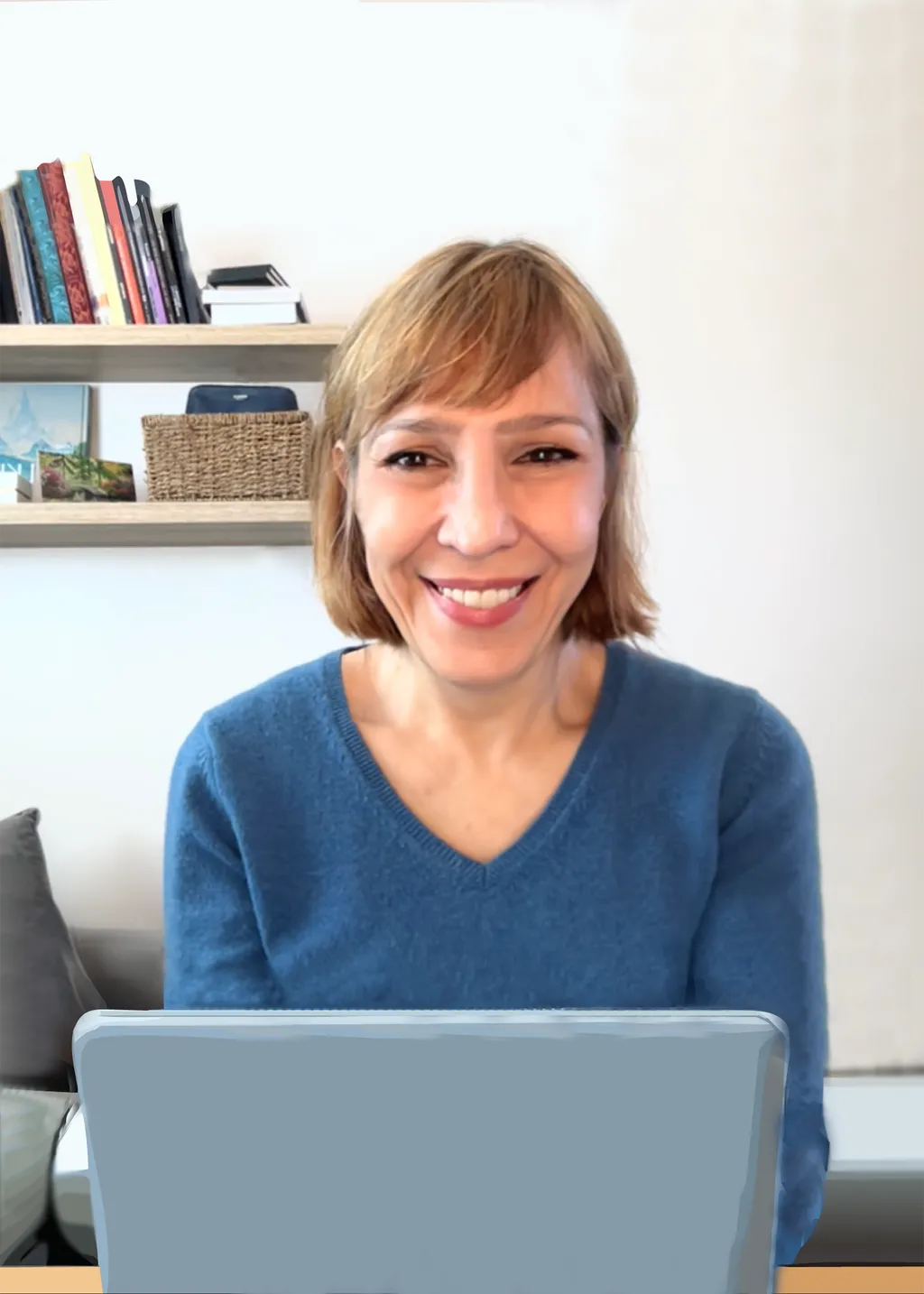 Woman with short blonde hair wearing blue jumper sits at laptop, smiling. White shelving with books visible behind her.