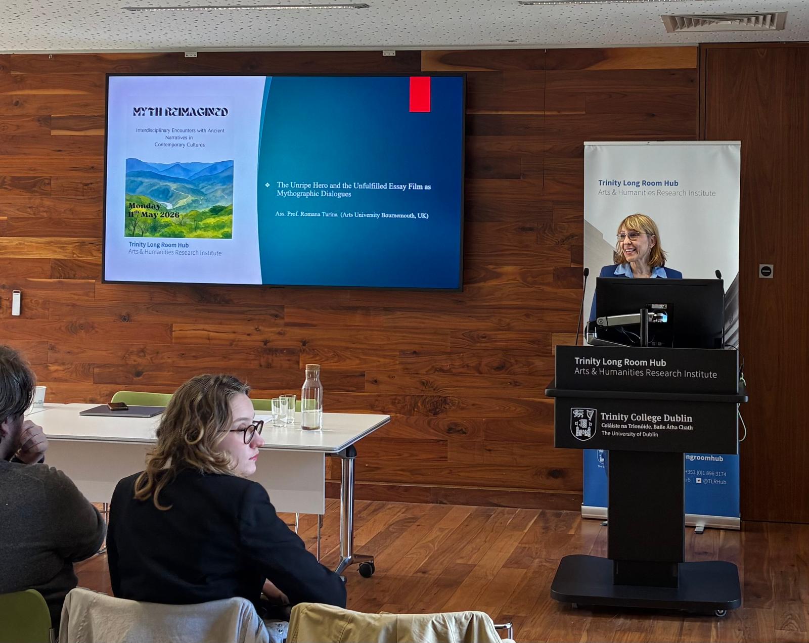 Associate Professor R Turina stands at a lectern delivering a presentation, displayed on a screen to the left of her.